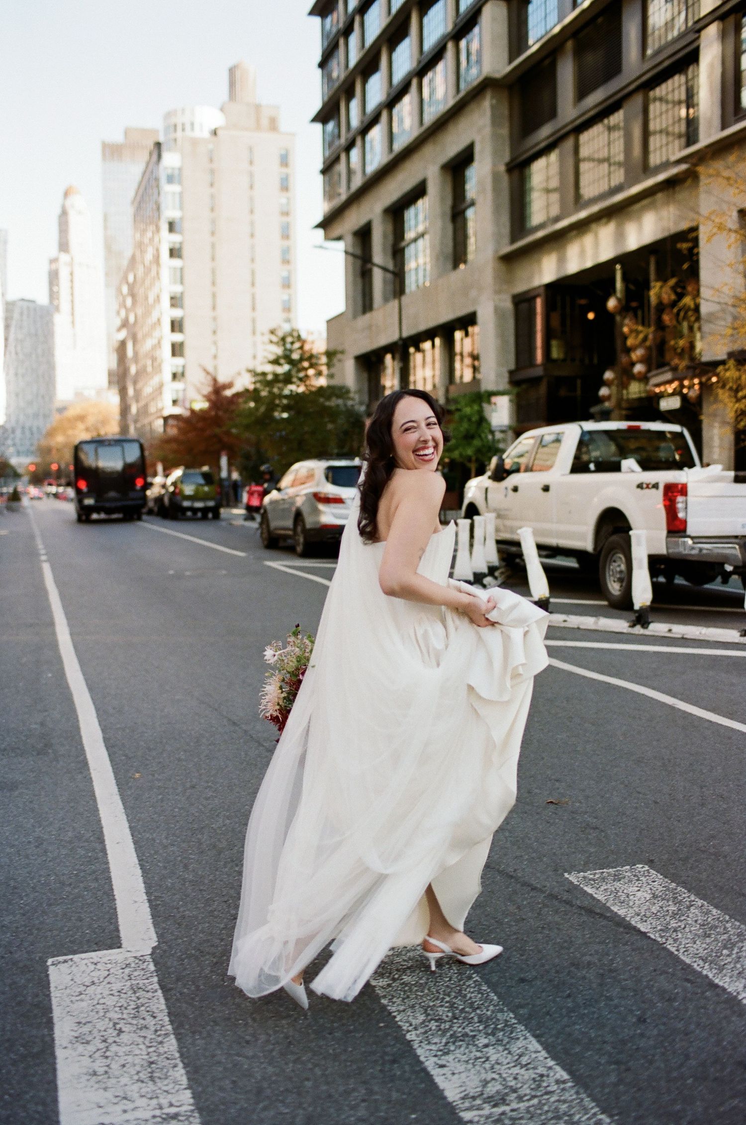 A person in a white wedding dress sits playfully on a city street with tall buildings in the background.