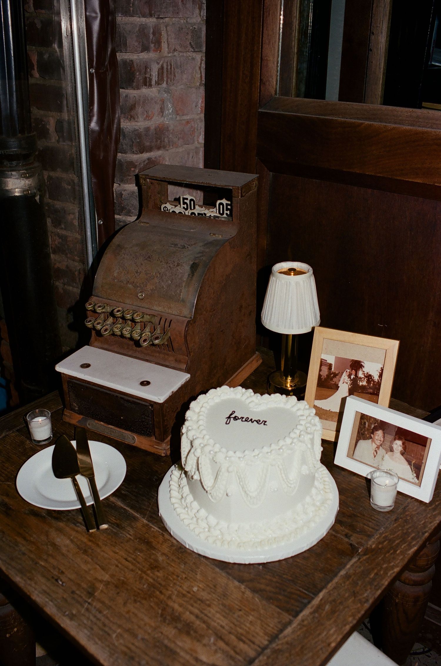 White wedding cake with frosted details sits on rustic wooden table next to framed photographs and table lamp.