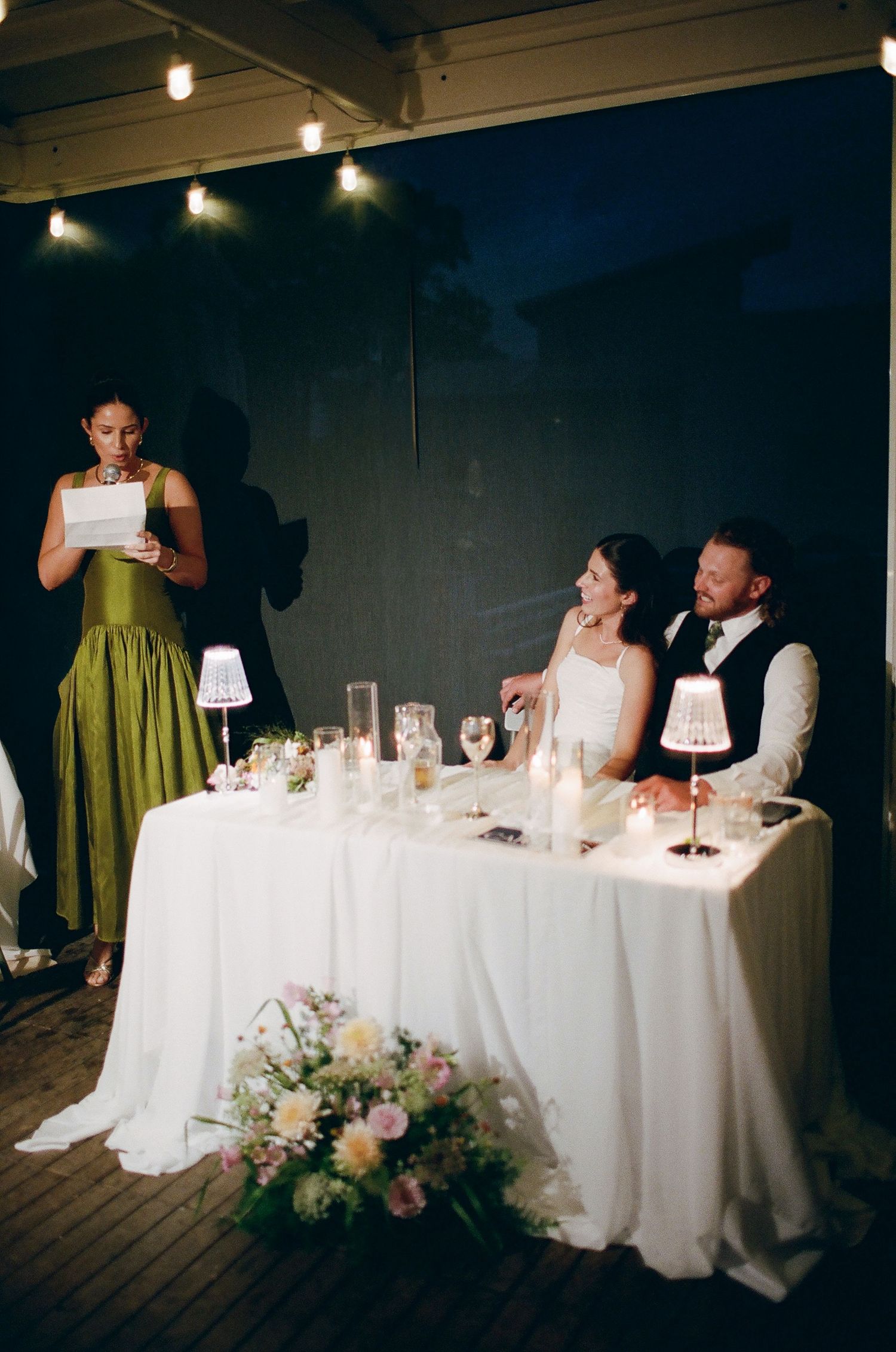 Bride and groom seated at sweetheart table while bridesmaid in green dress delivers wedding speech at evening reception.