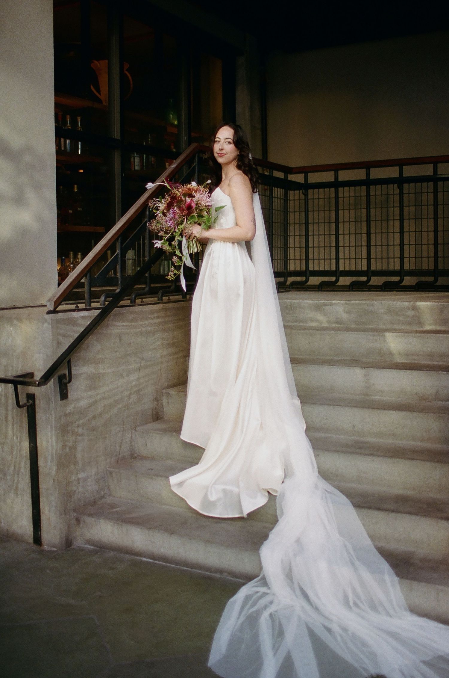 A bride in a flowing white wedding dress with a long train poses on concrete steps in an elegant architectural setting.