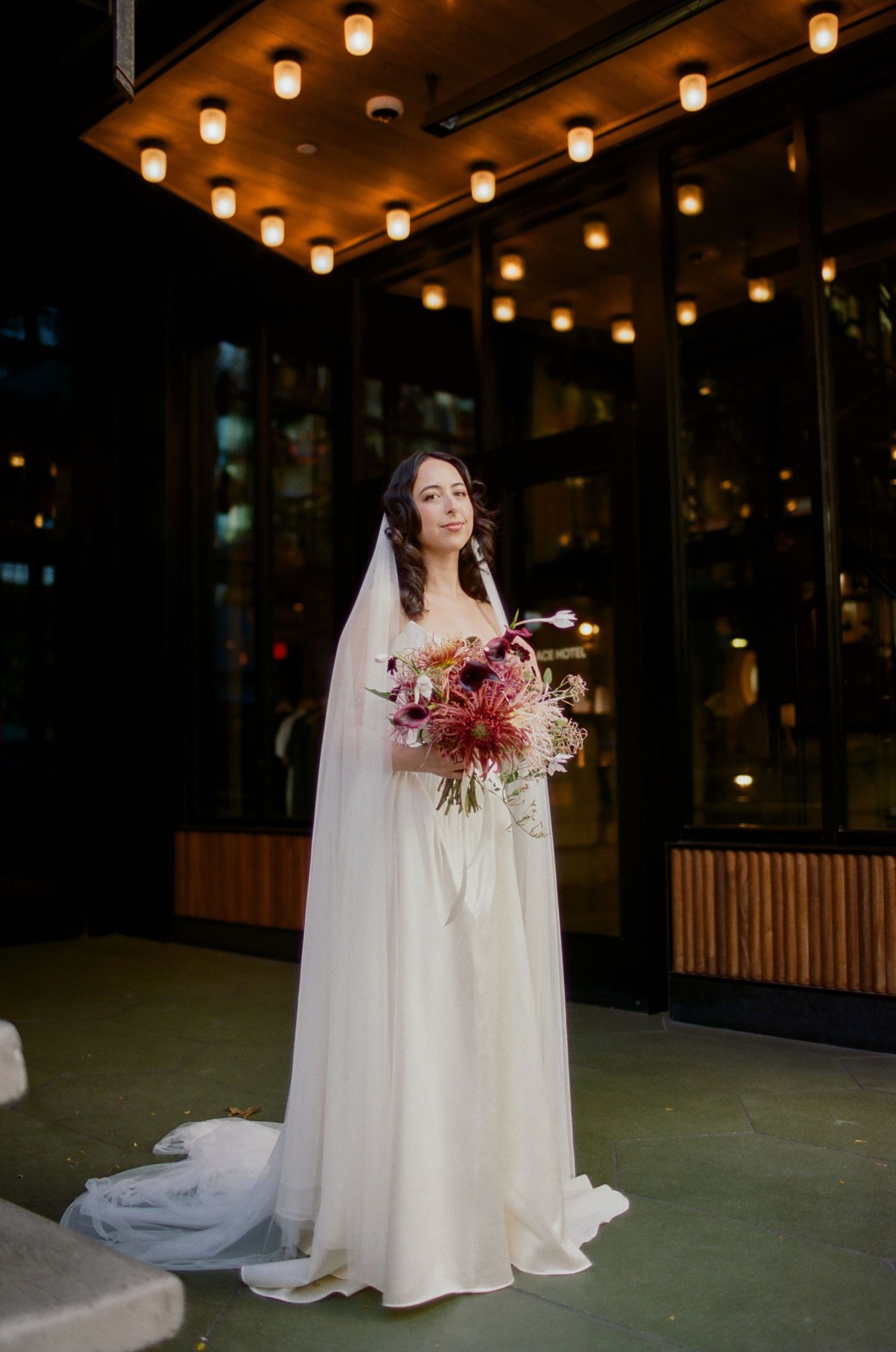 A bride in a flowing white wedding dress holds a vibrant red and pink bouquet under warm indoor lighting.