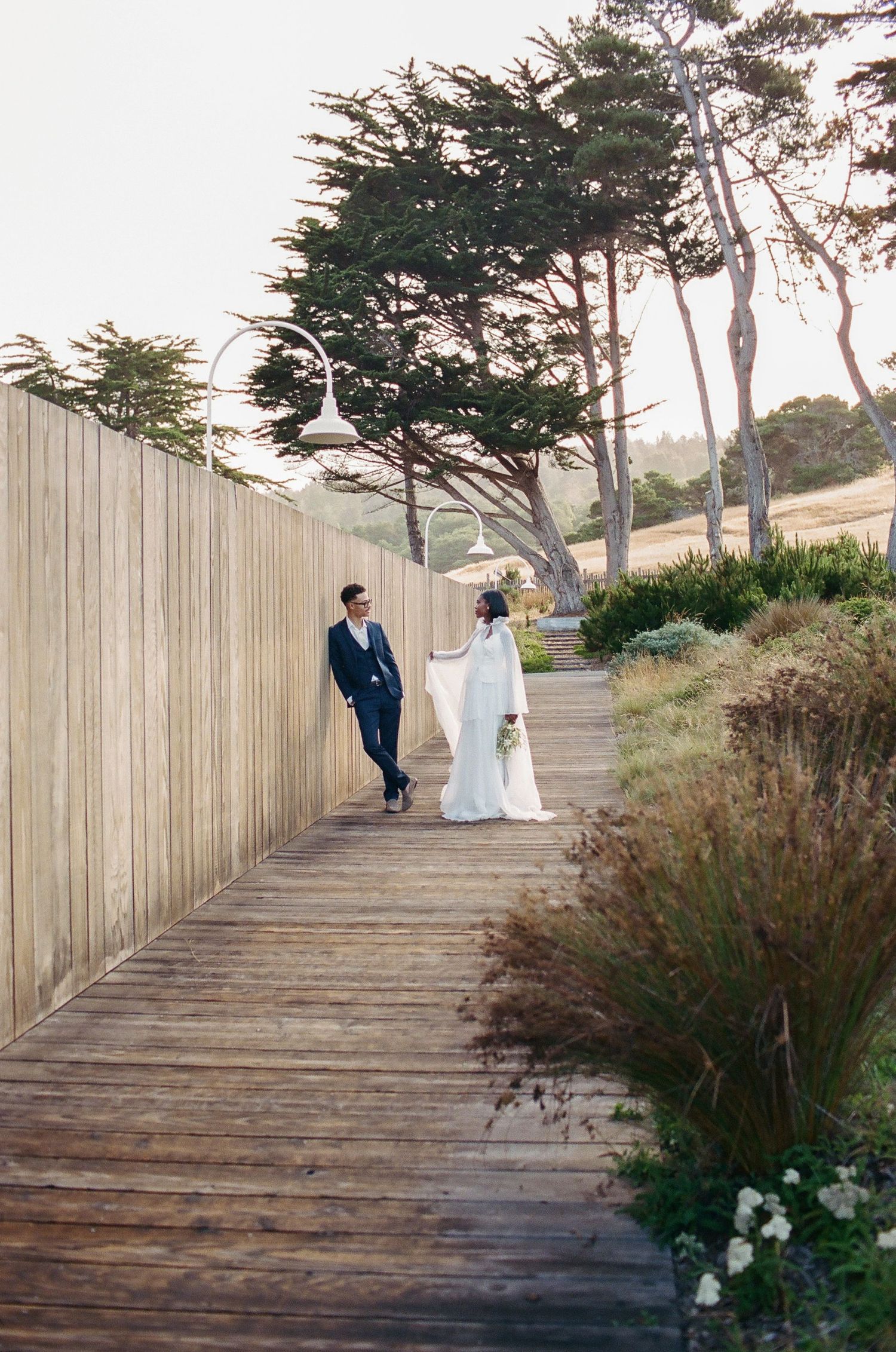 Bride and groom share a romantic moment on a wooden boardwalk path surrounded by windswept cypress trees.