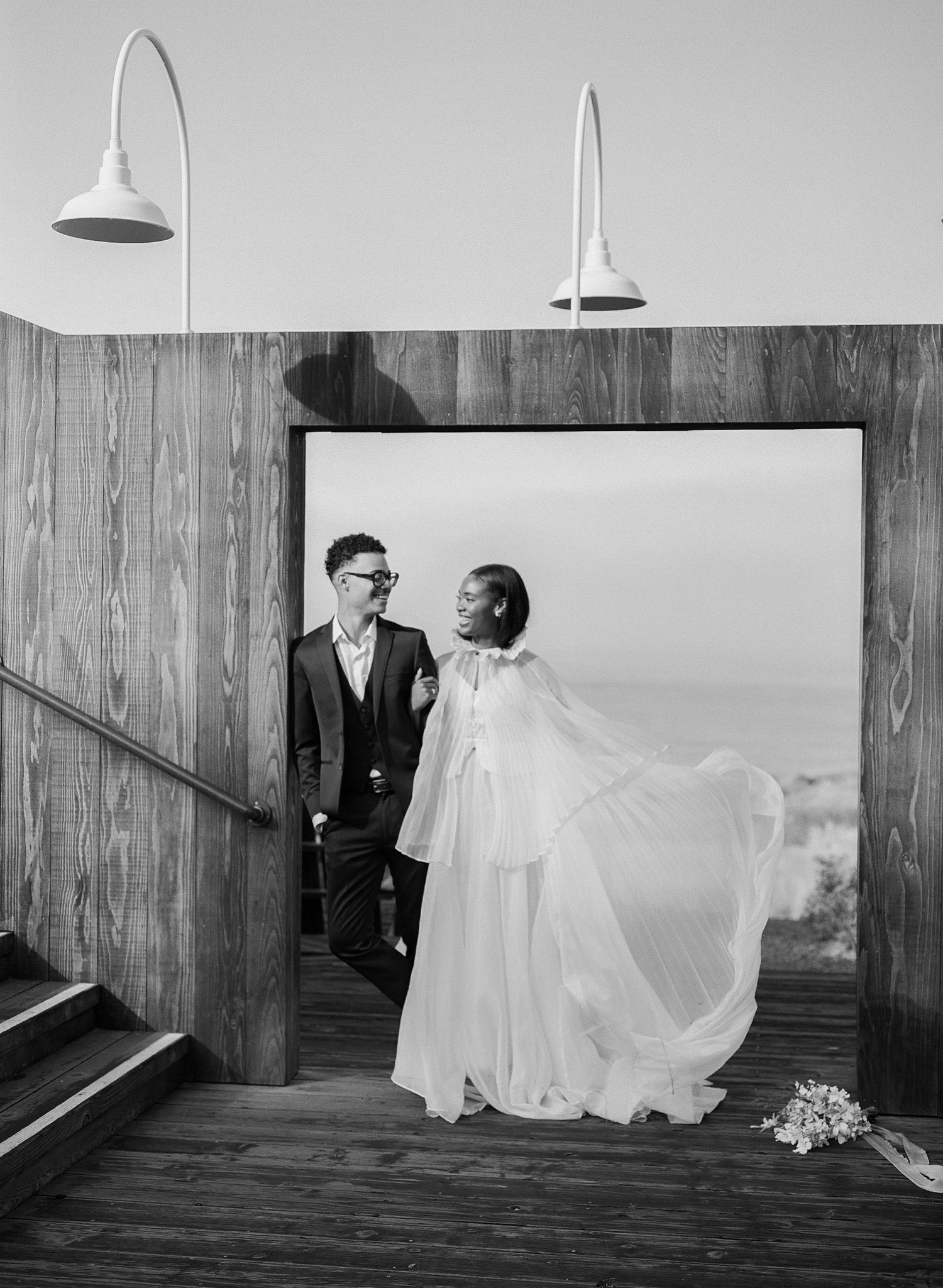 Black and white photo of a couple in wedding attire standing in a wooden doorway, bride's cape flowing in the wind.