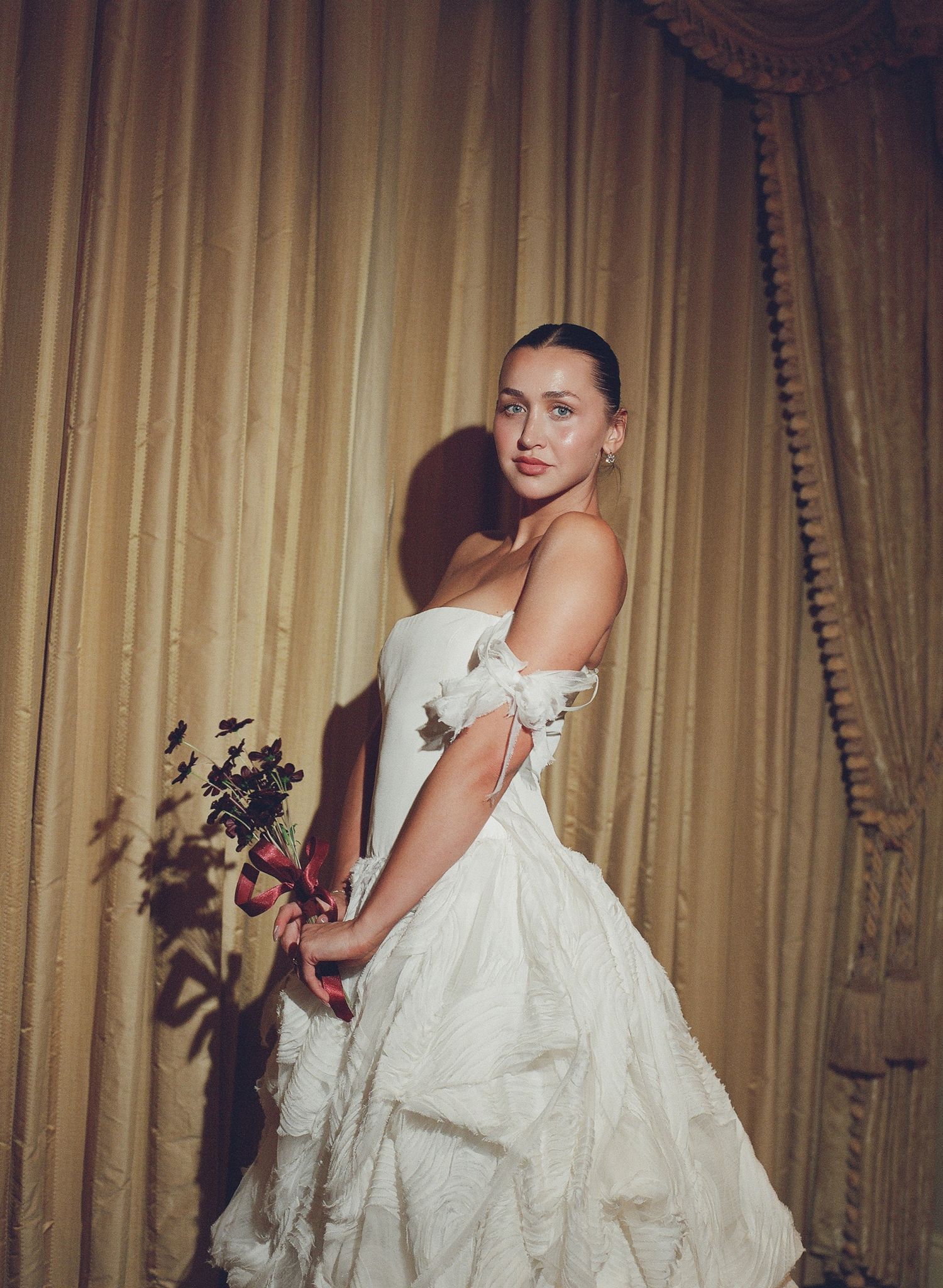 Woman in white off-shoulder ball gown holding dark floral bouquet, posing before gold curtain backdrop.
