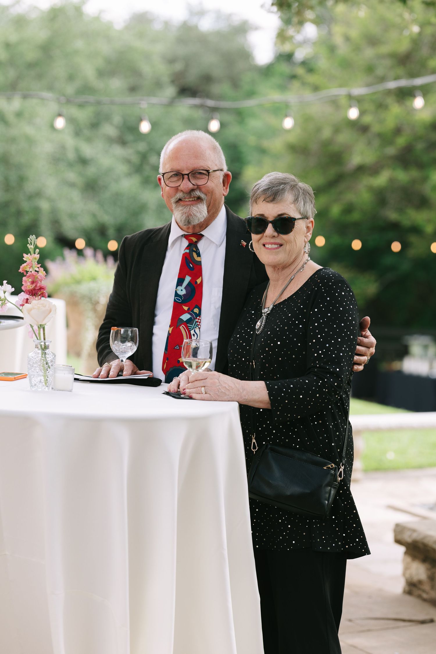 Elegant couple enjoying drinks at a cocktail hour by a white standing table with string lights in the background.