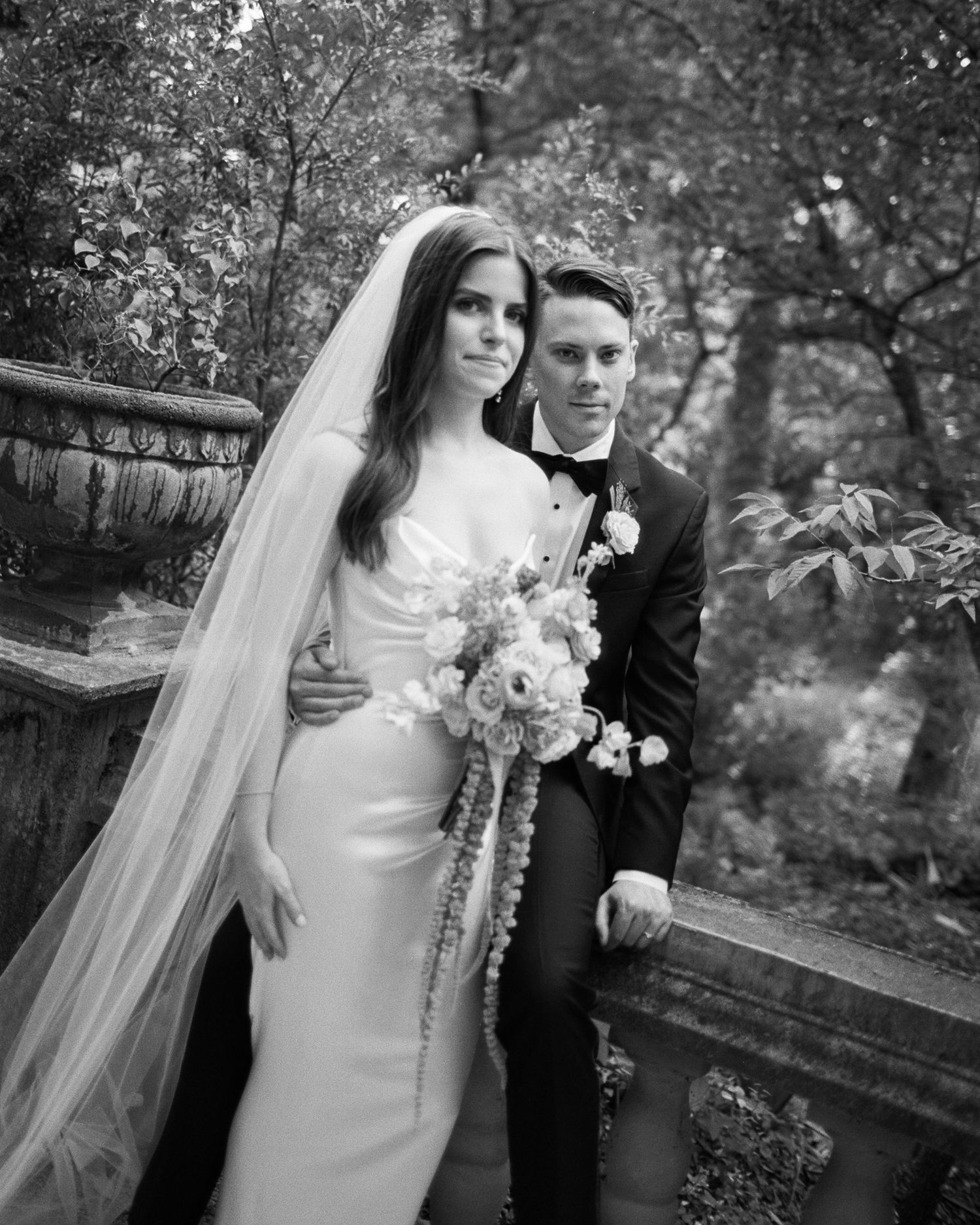 A wedding couple poses together in a garden setting holding an elegant white floral bouquet.