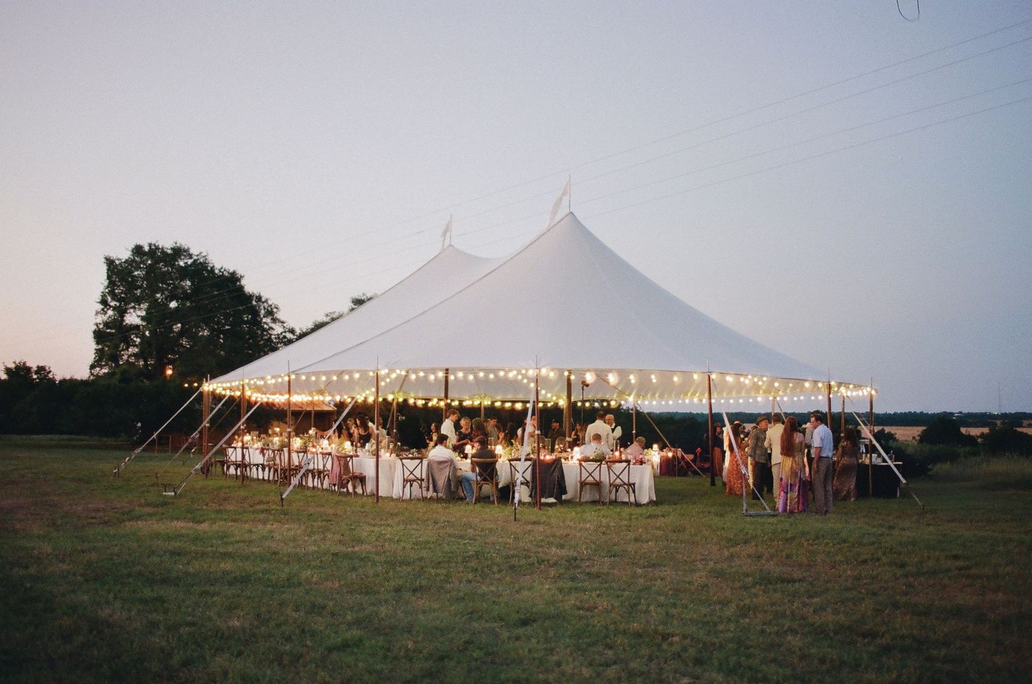 Elegant outdoor wedding reception under a large white sailcloth tent with string lights at dusk on a green field.