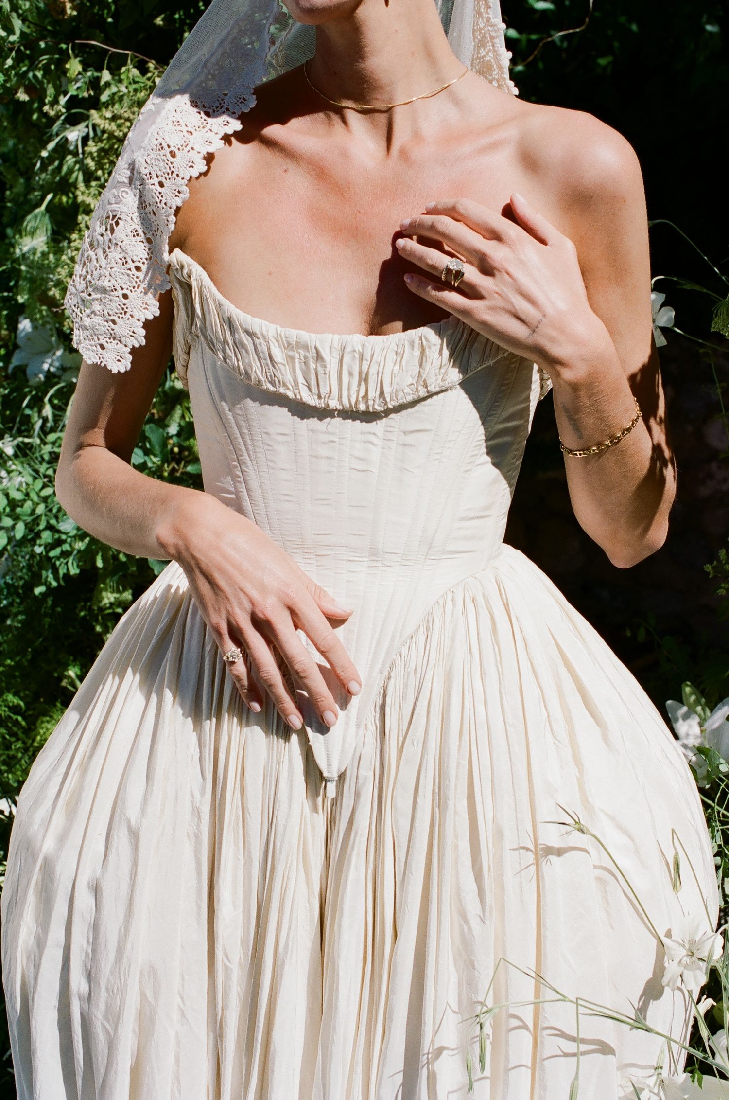 Bride in ivory off-shoulder pleated wedding gown with lace sleeve detail and veil, posing outdoors among greenery.