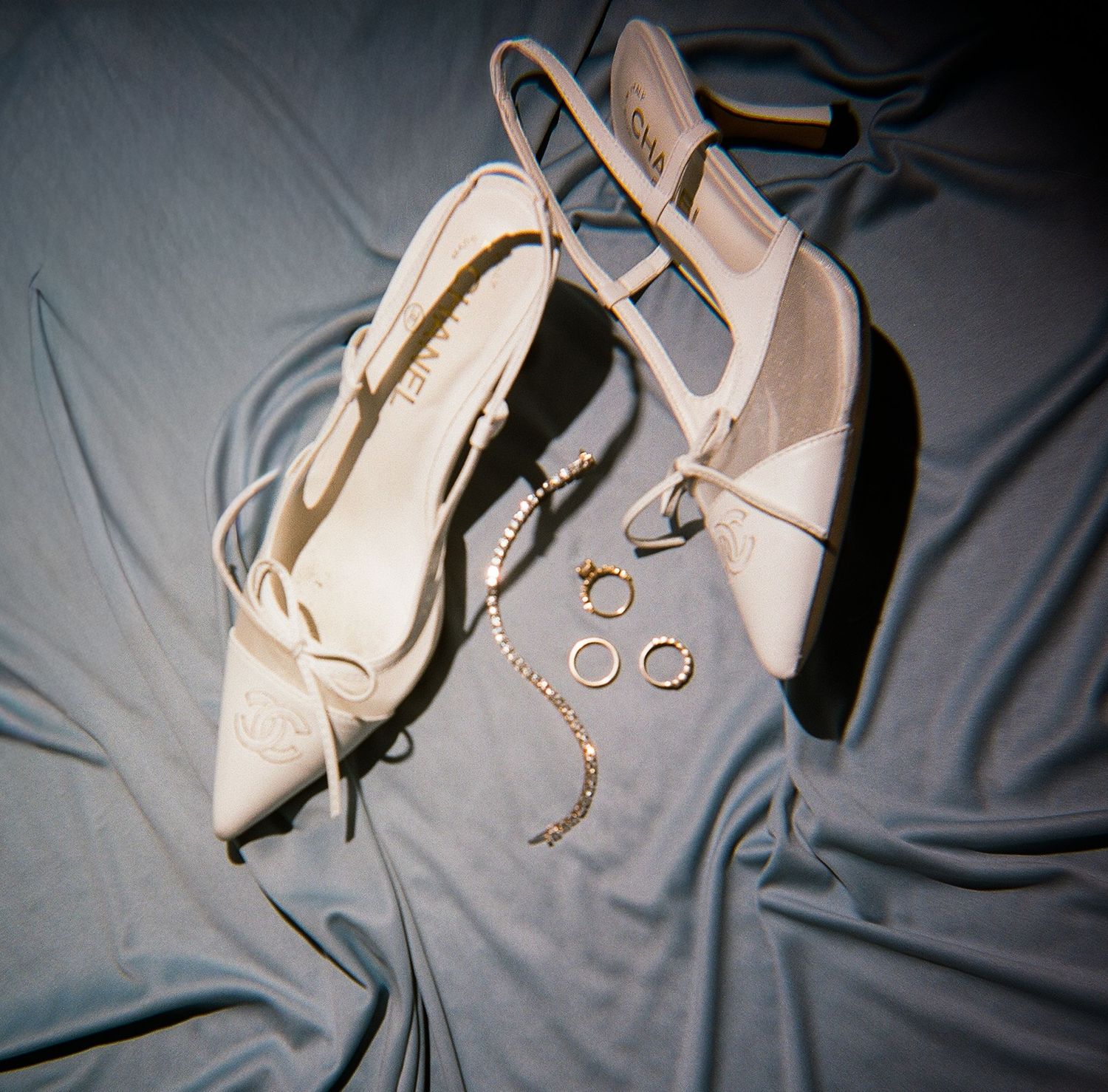 White satin slingback heels displayed on blue silk fabric alongside gold hoop earrings and a delicate pearl bracelet.