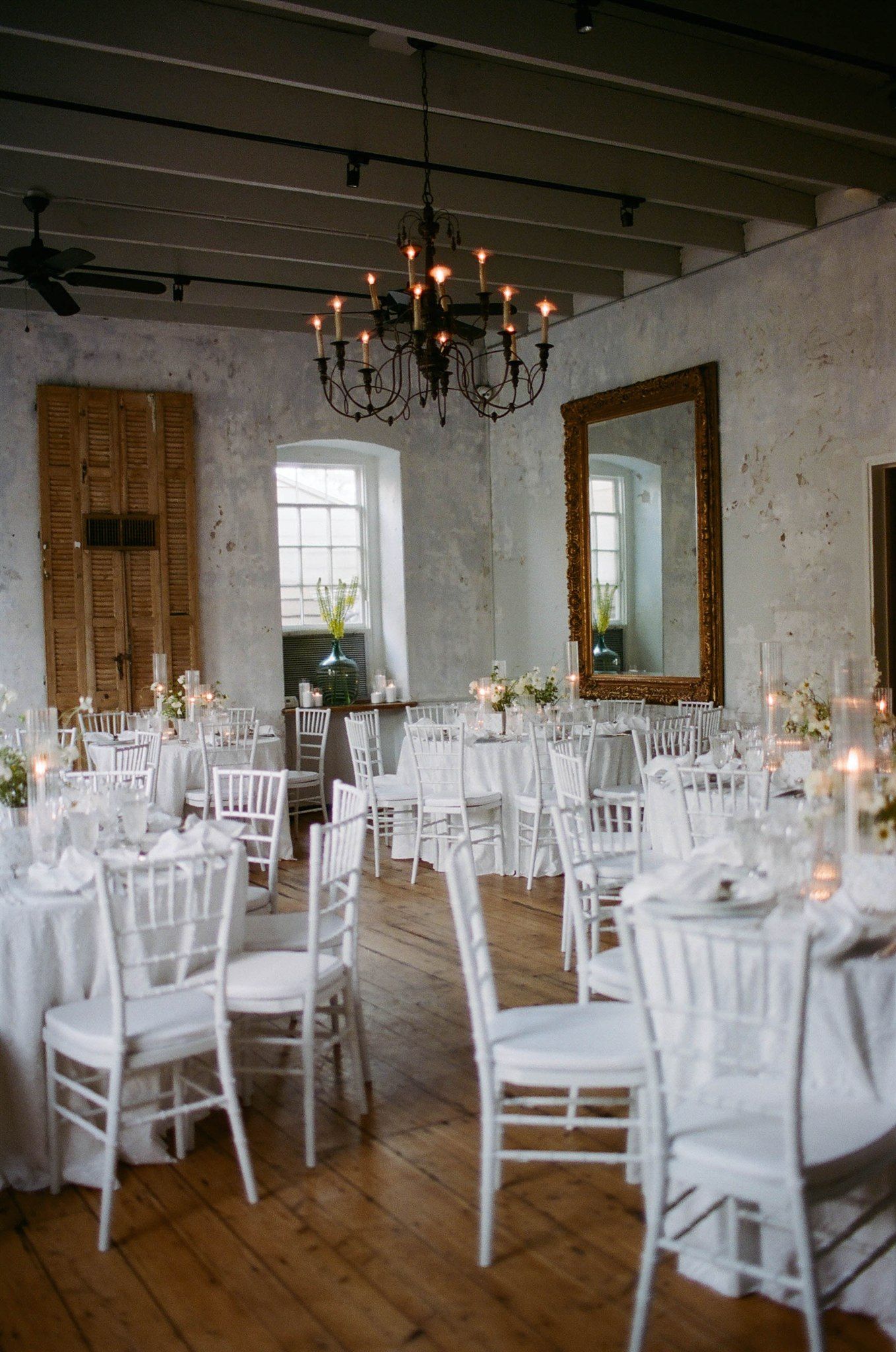 Rustic white reception room with chandelier and wooden floors set with round tables and chairs.