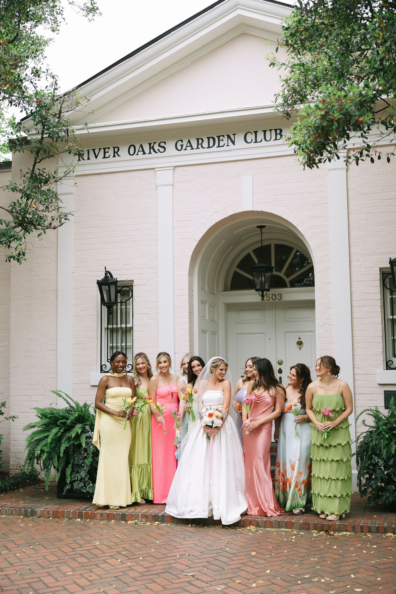 Wedding party lineup in yellow, pink and green dresses outside white colonial building.