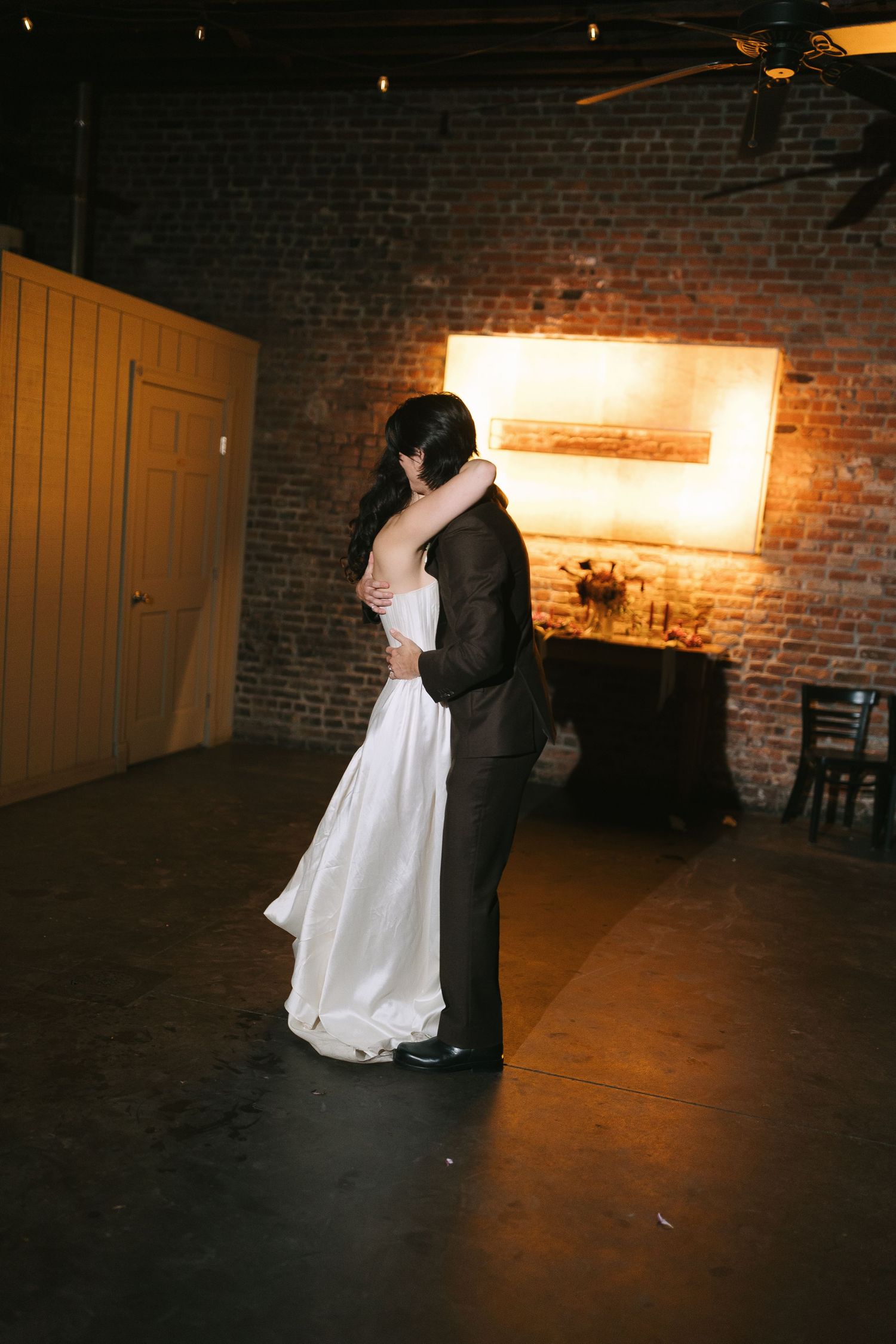Wedding couple dances in rustic brick-walled venue with warm lighting.