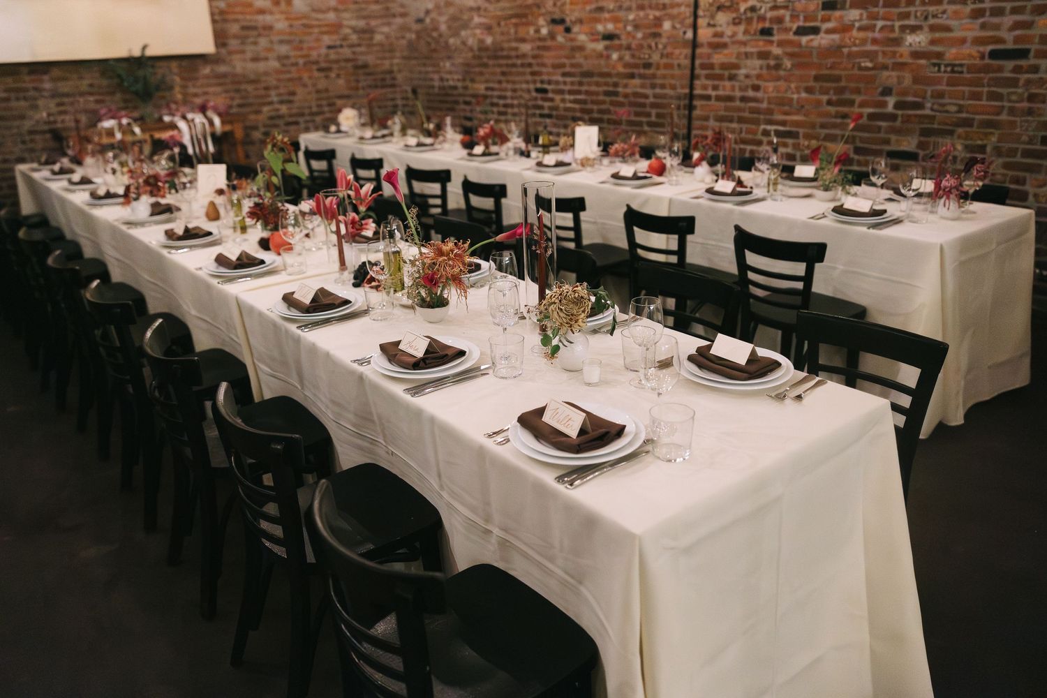 Formal dining table setup with black chairs and floral centerpieces against exposed brick wall.