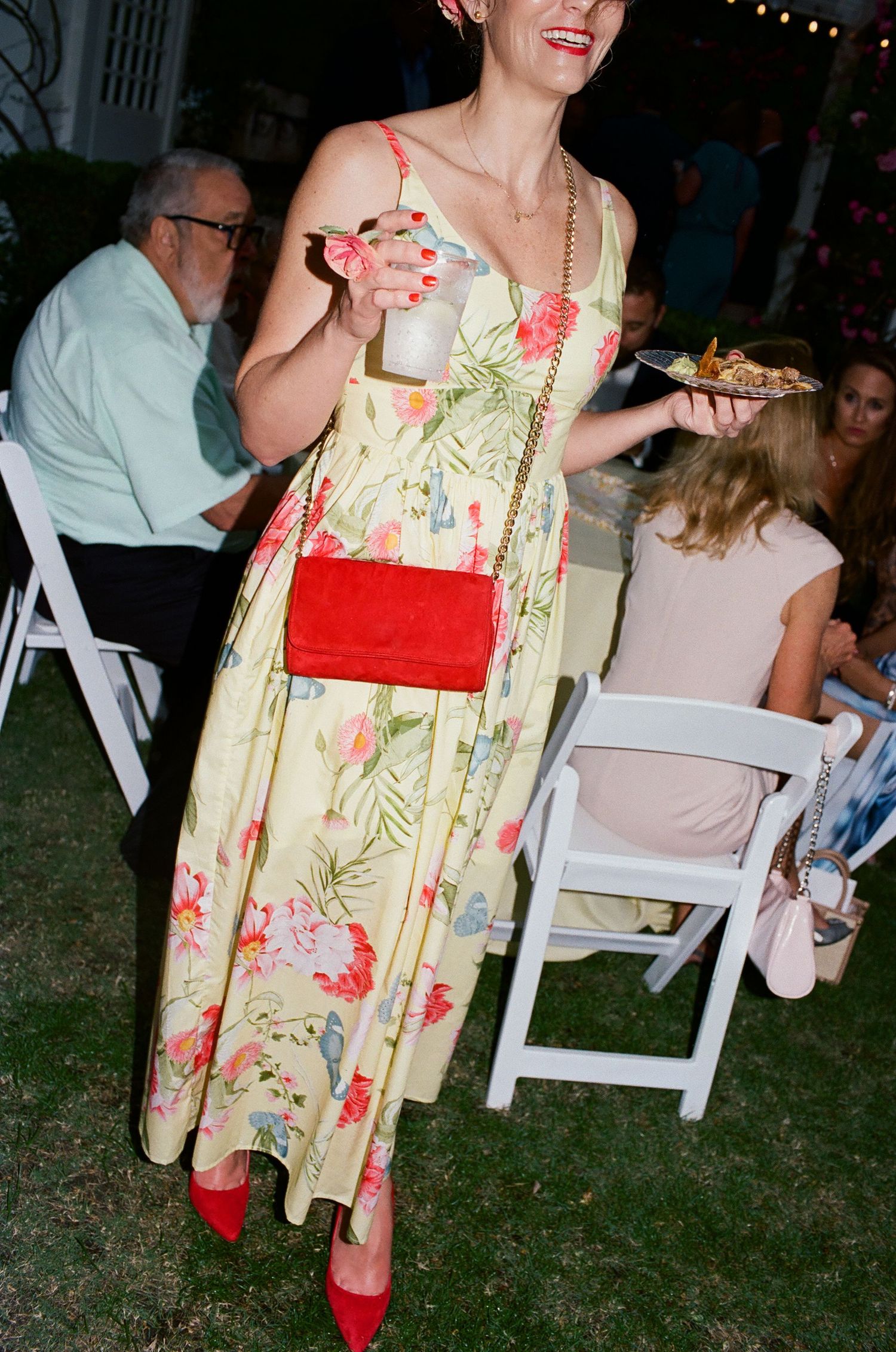 Wedding guests in floral dresses enjoy an outdoor evening reception with white chairs.