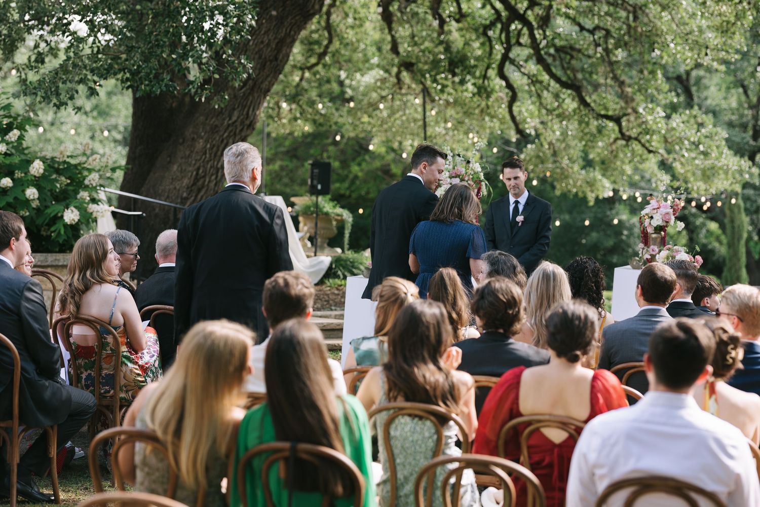 Rows of wedding guests seated outdoors for a garden ceremony under tree branches.