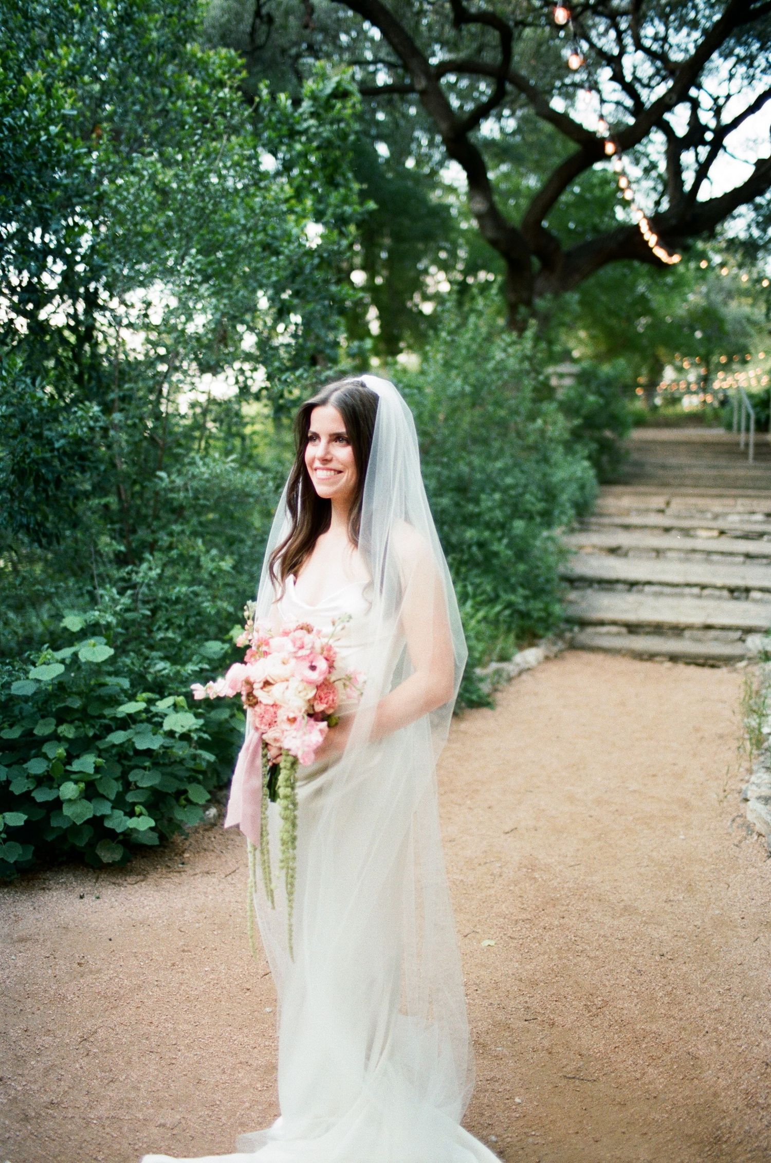 Bride in white gown holds pink bouquet on garden path surrounded by trees.