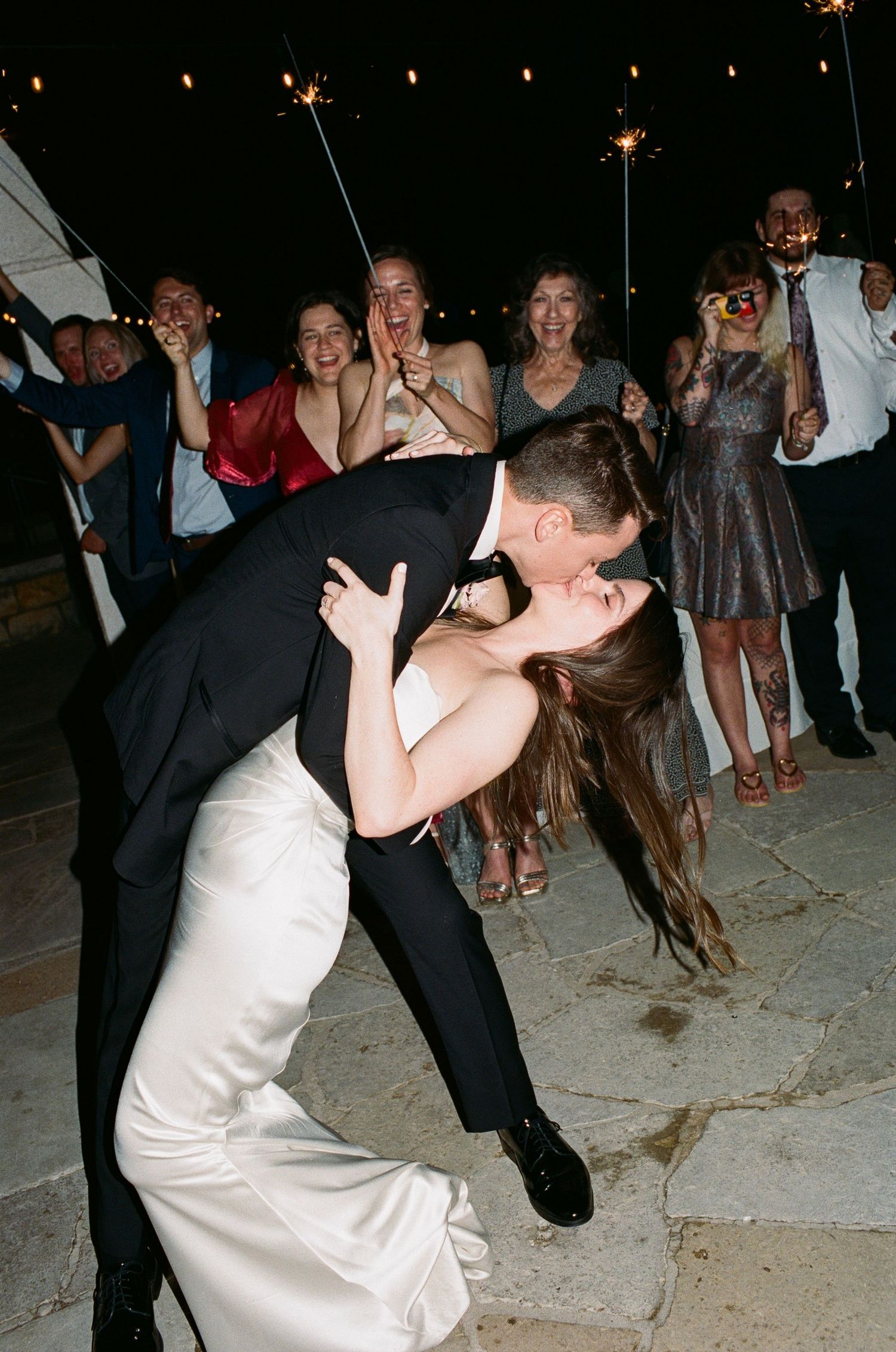 A romantic dip kiss on the dance floor while wedding guests hold sparklers in the background.