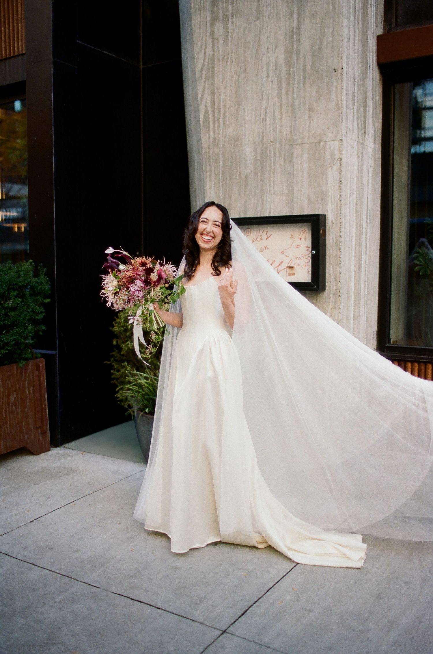 A bride in a flowing white wedding dress twirls and poses on a concrete sidewalk outside a modern building.