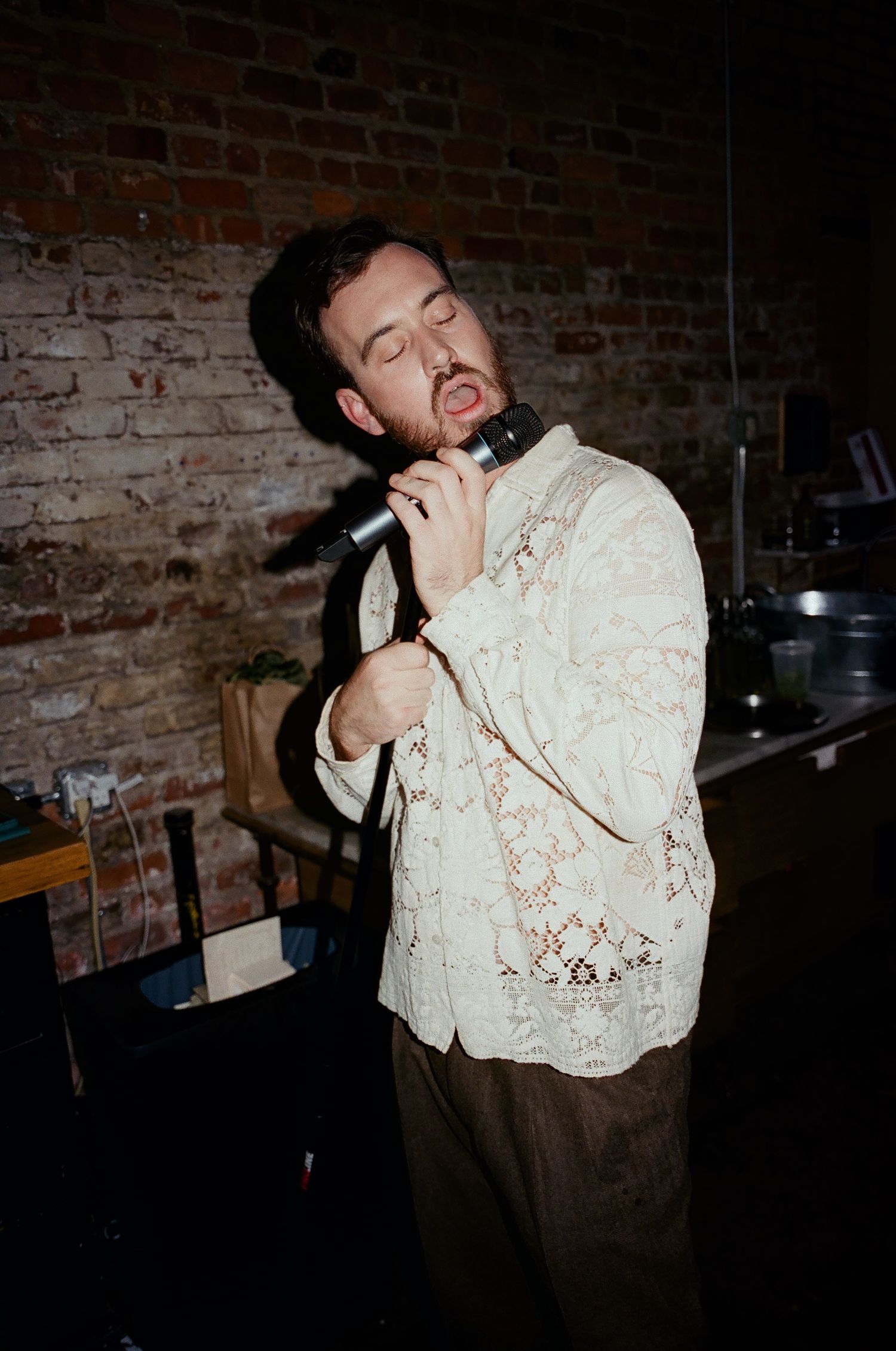 A young man with a beard sings passionately into a microphone wearing a cream lace sweater against a brick wall.