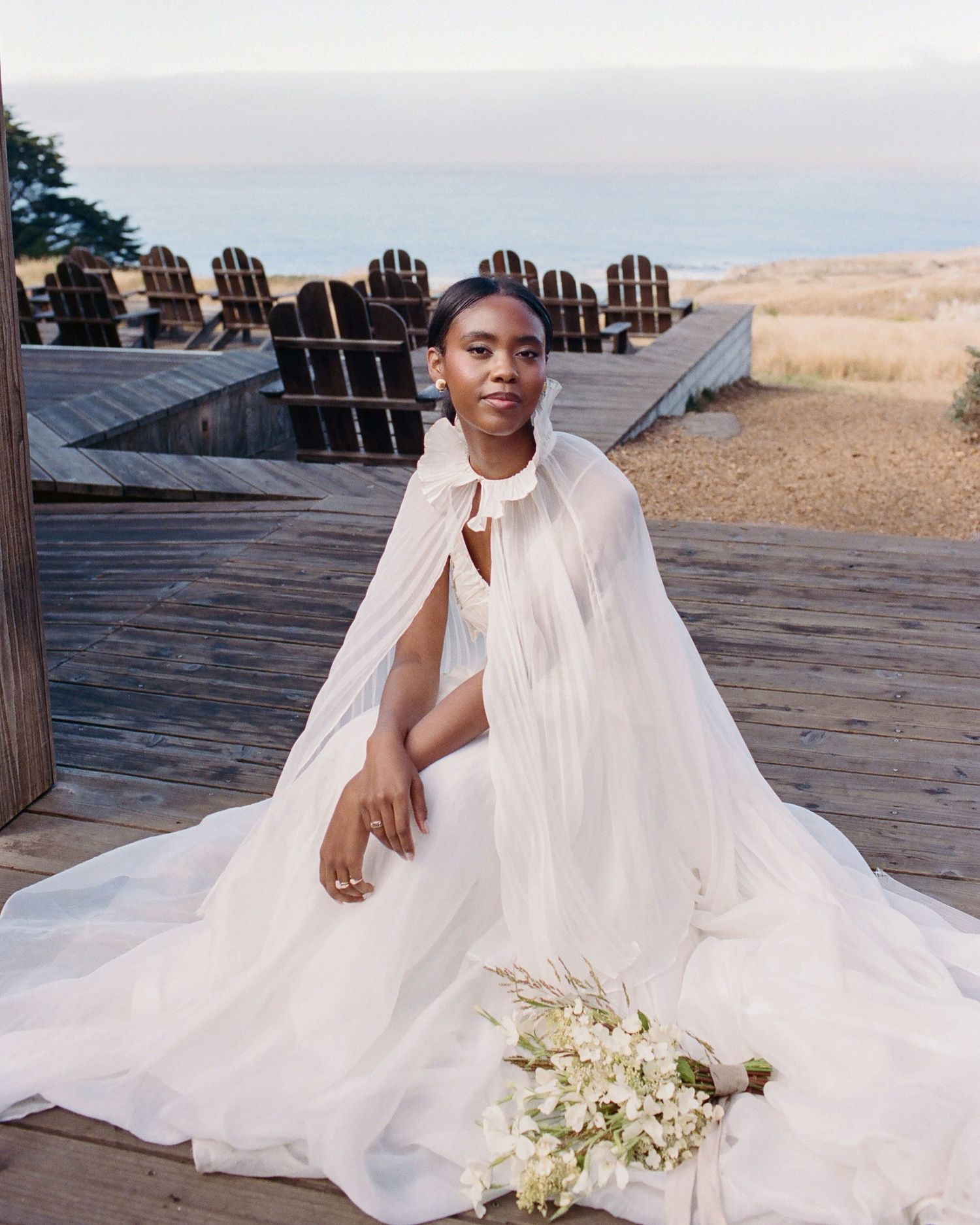 A Black bride in a white flowing gown with a chiffon cape holds a bouquet, seated on a wooden deck overlooking the ocean.