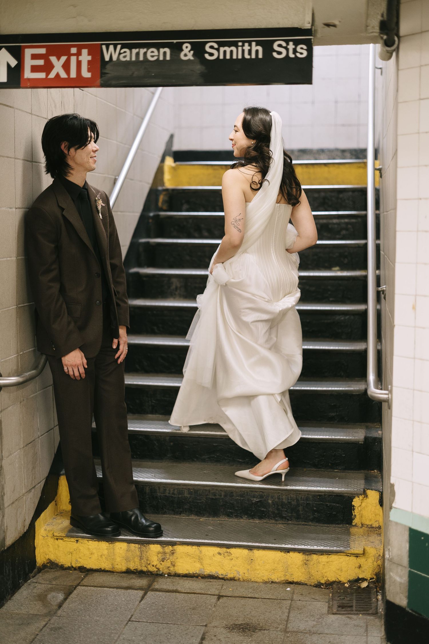 Bride and groom share playful moment on subway station steps under exit sign.