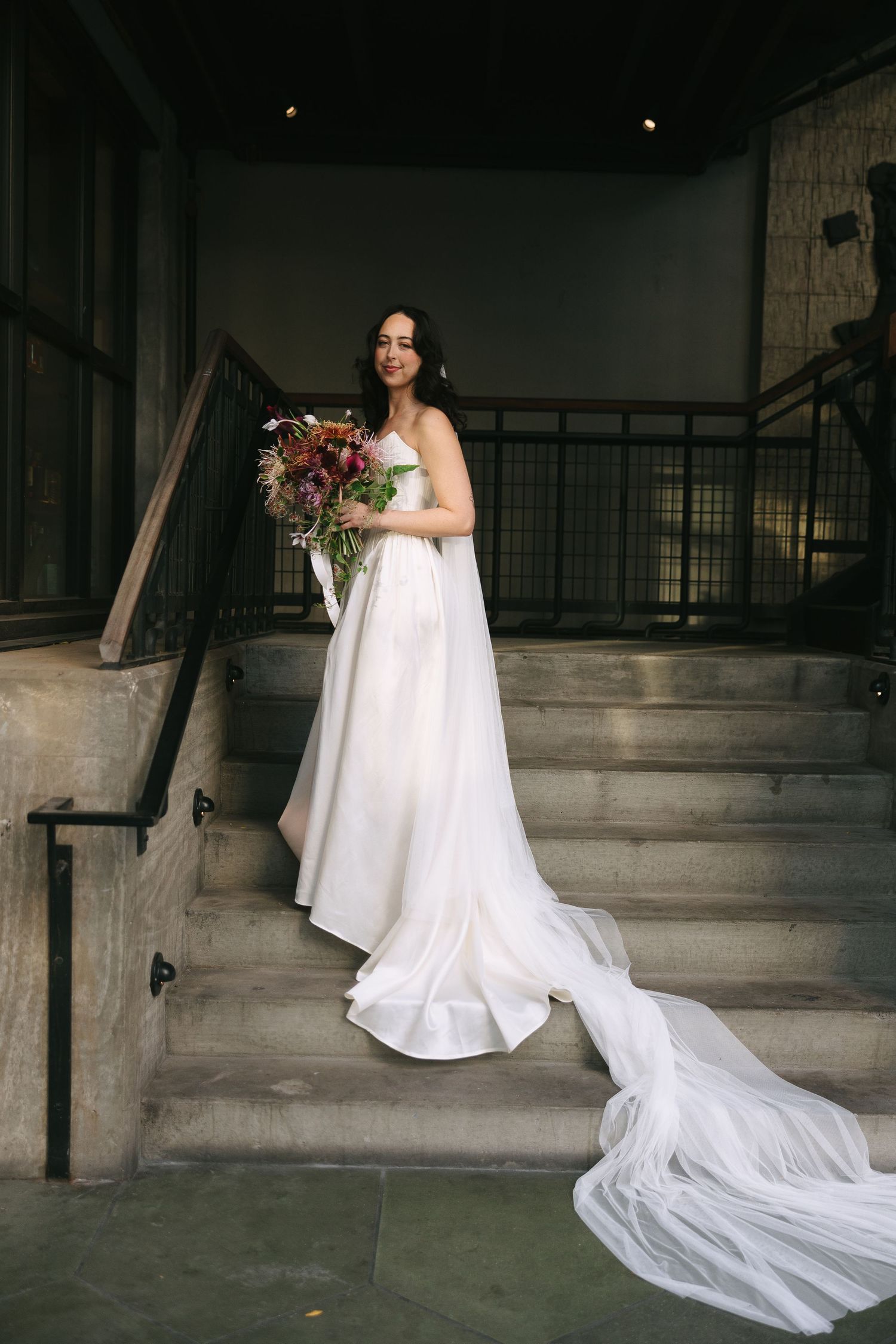 An elegant white wedding dress with long train photographed on industrial concrete stairs.