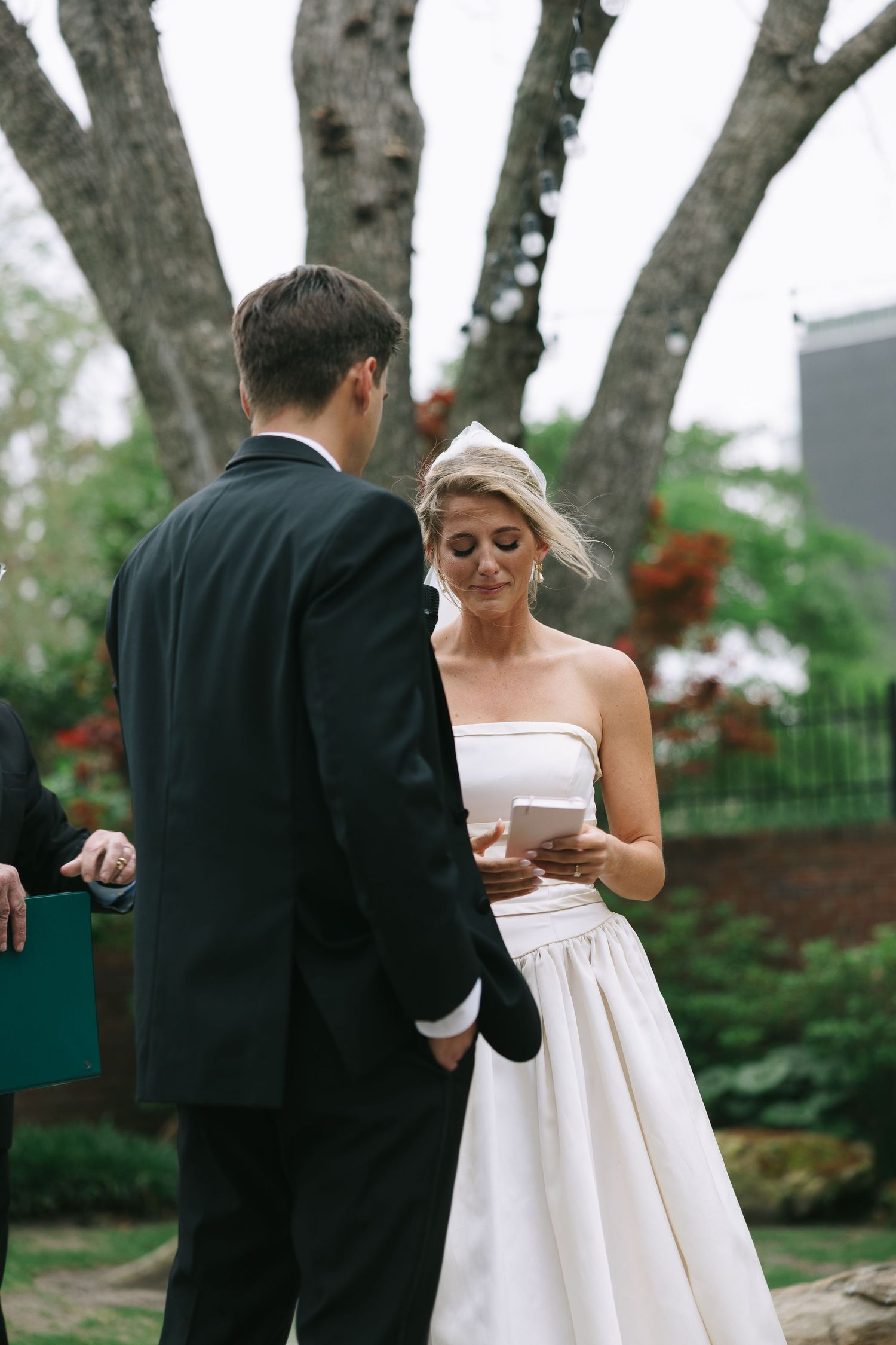 Bride in white strapless gown reading during outdoor wedding ceremony.