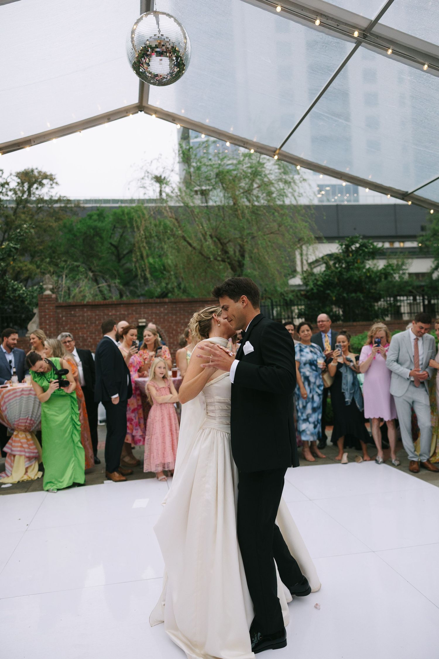 Guests watch as a couple dances under a white tent with a disco ball overhead.