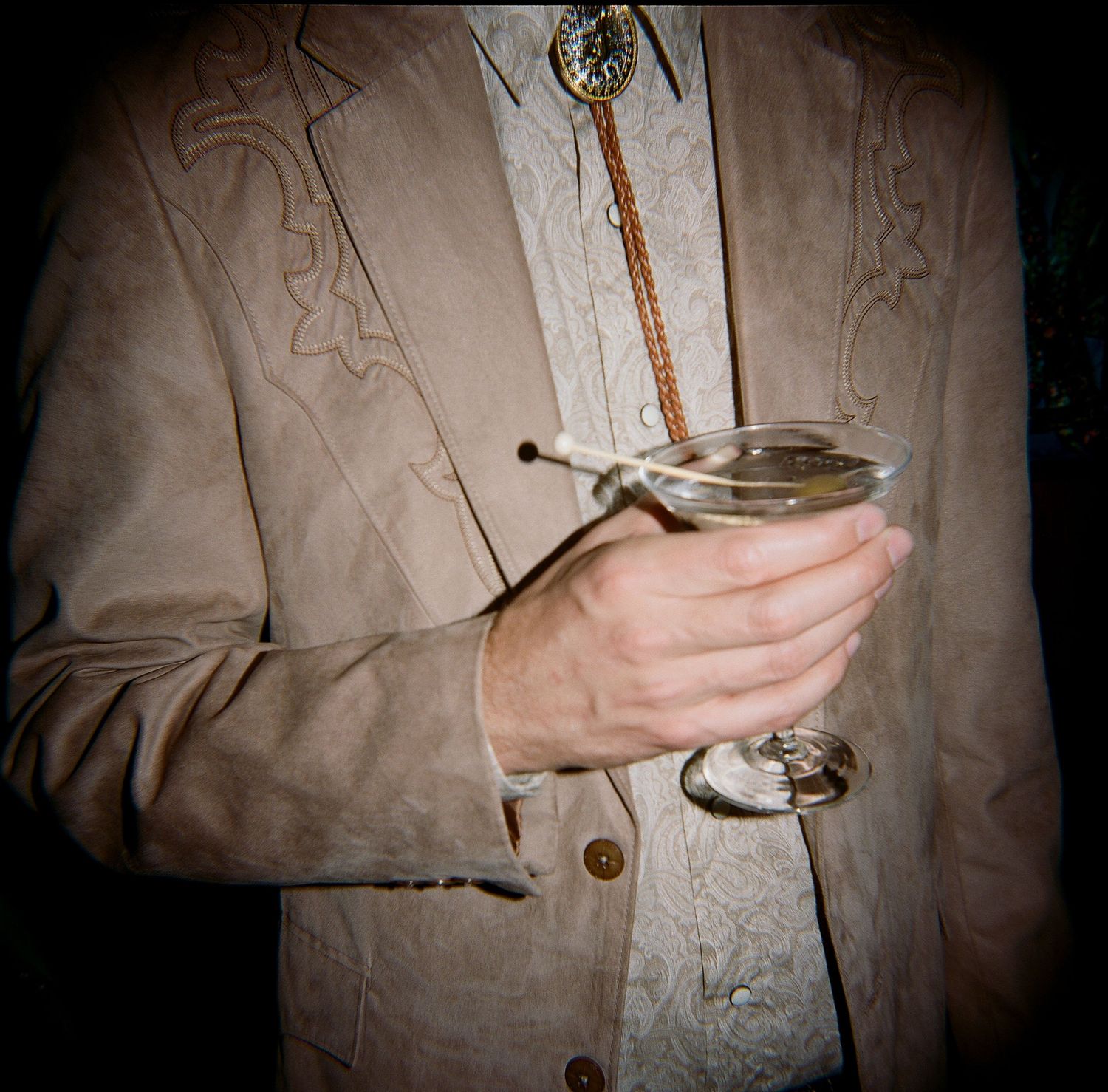 Man in tan suede western jacket holding a martini glass with a bolo tie, close-up shot.