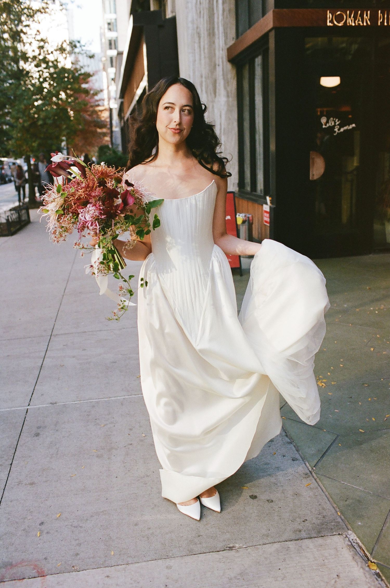 A person in a flowing white wedding dress holds a vibrant autumn bouquet while walking on a city sidewalk.