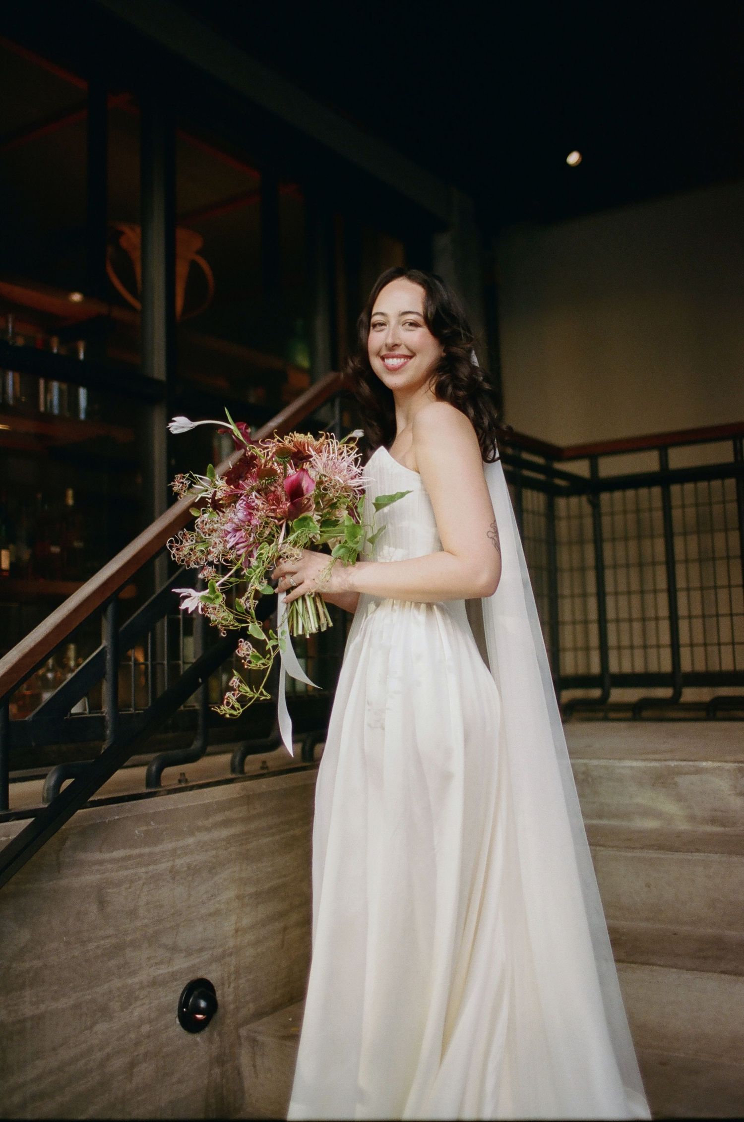 A bride in a flowing white wedding dress holds a vibrant burgundy and pink bouquet in an industrial venue.