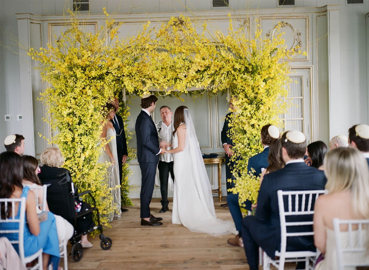 Wedding ceremony setup with bright yellow floral arch and white chairs in an indoor venue.