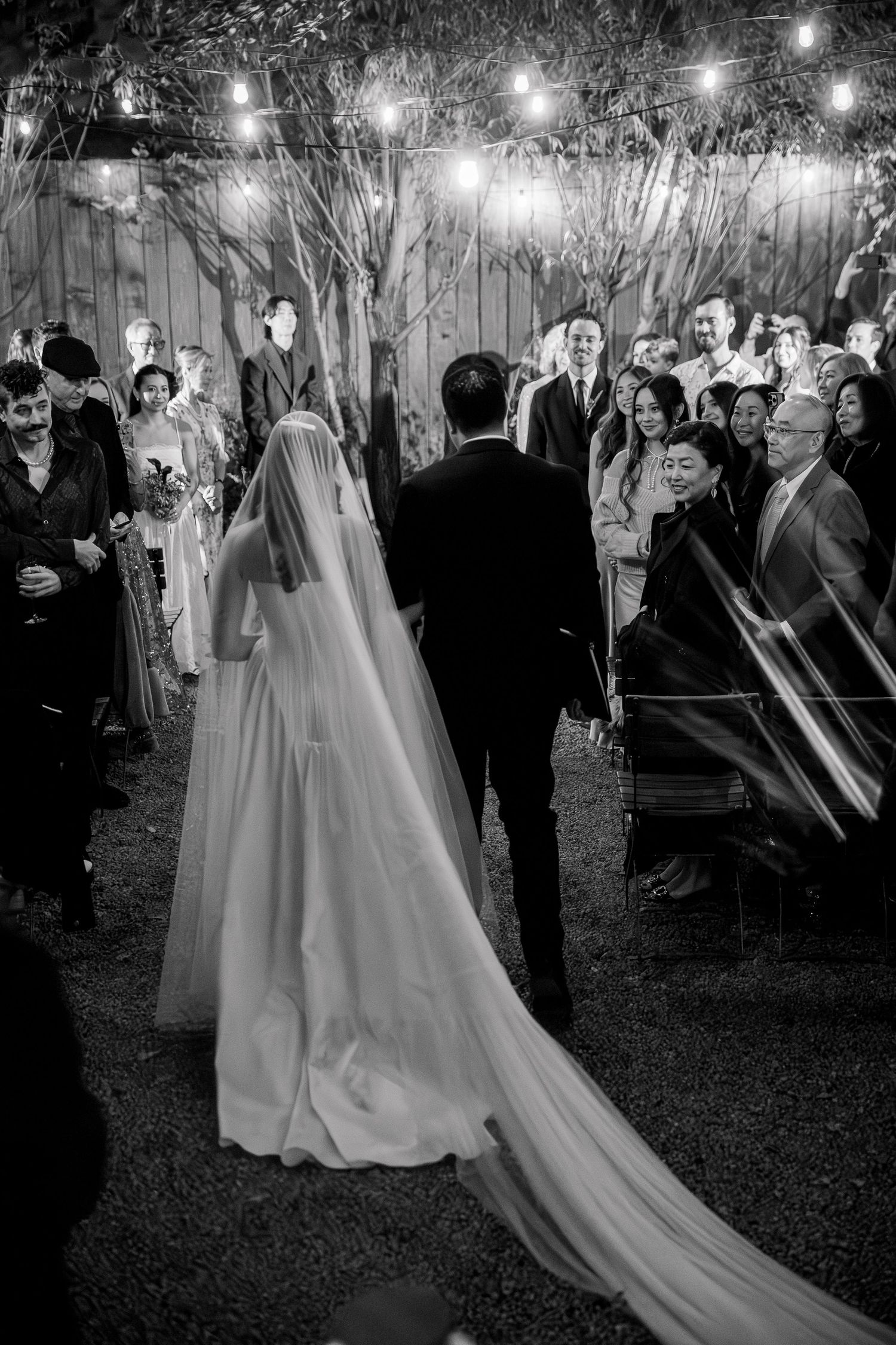 A bride in a flowing white gown walks through an outdoor evening reception with string lights overhead.