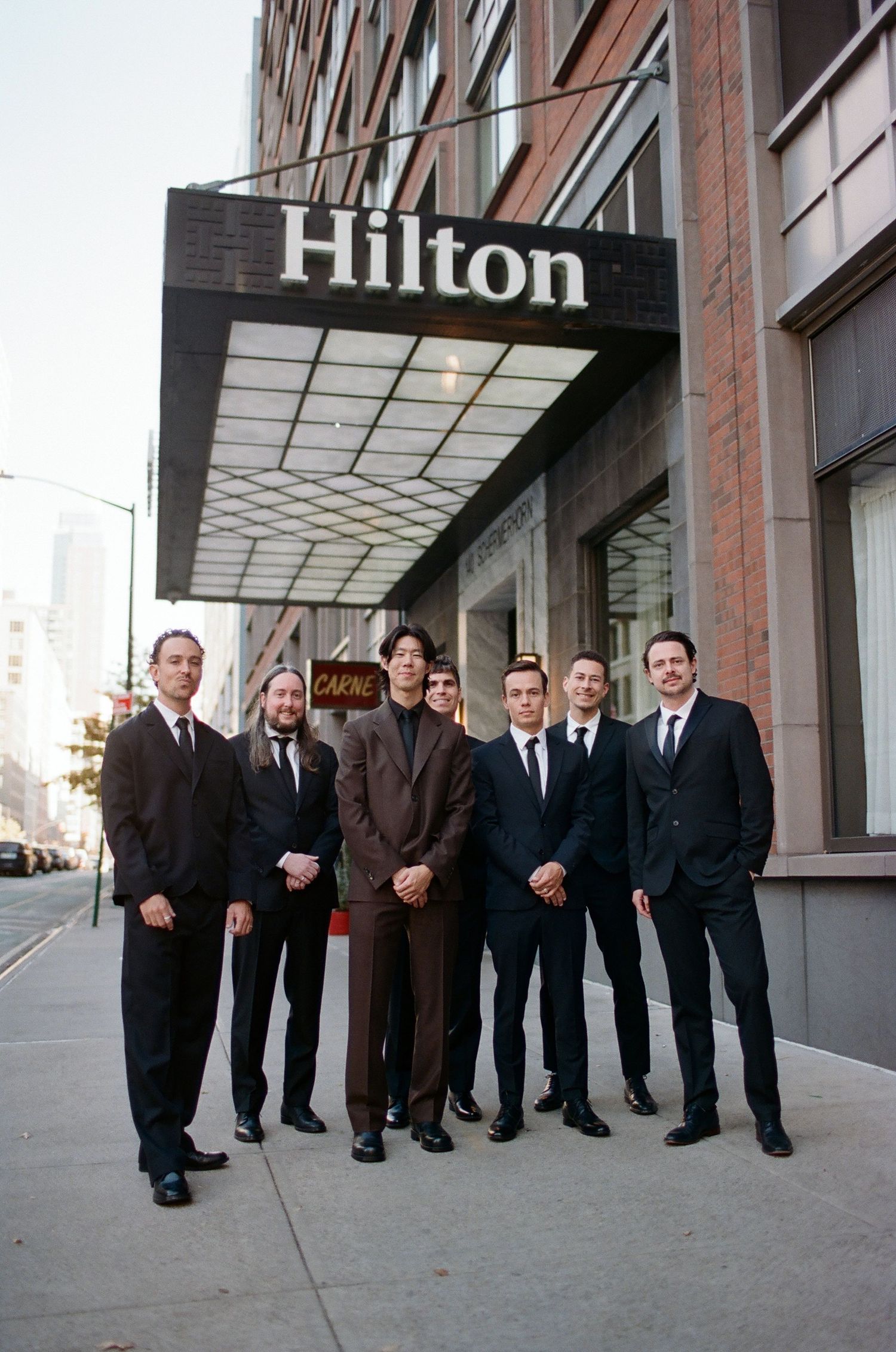A group of formally dressed men in dark suits standing in a line outside a Hilton hotel entrance.