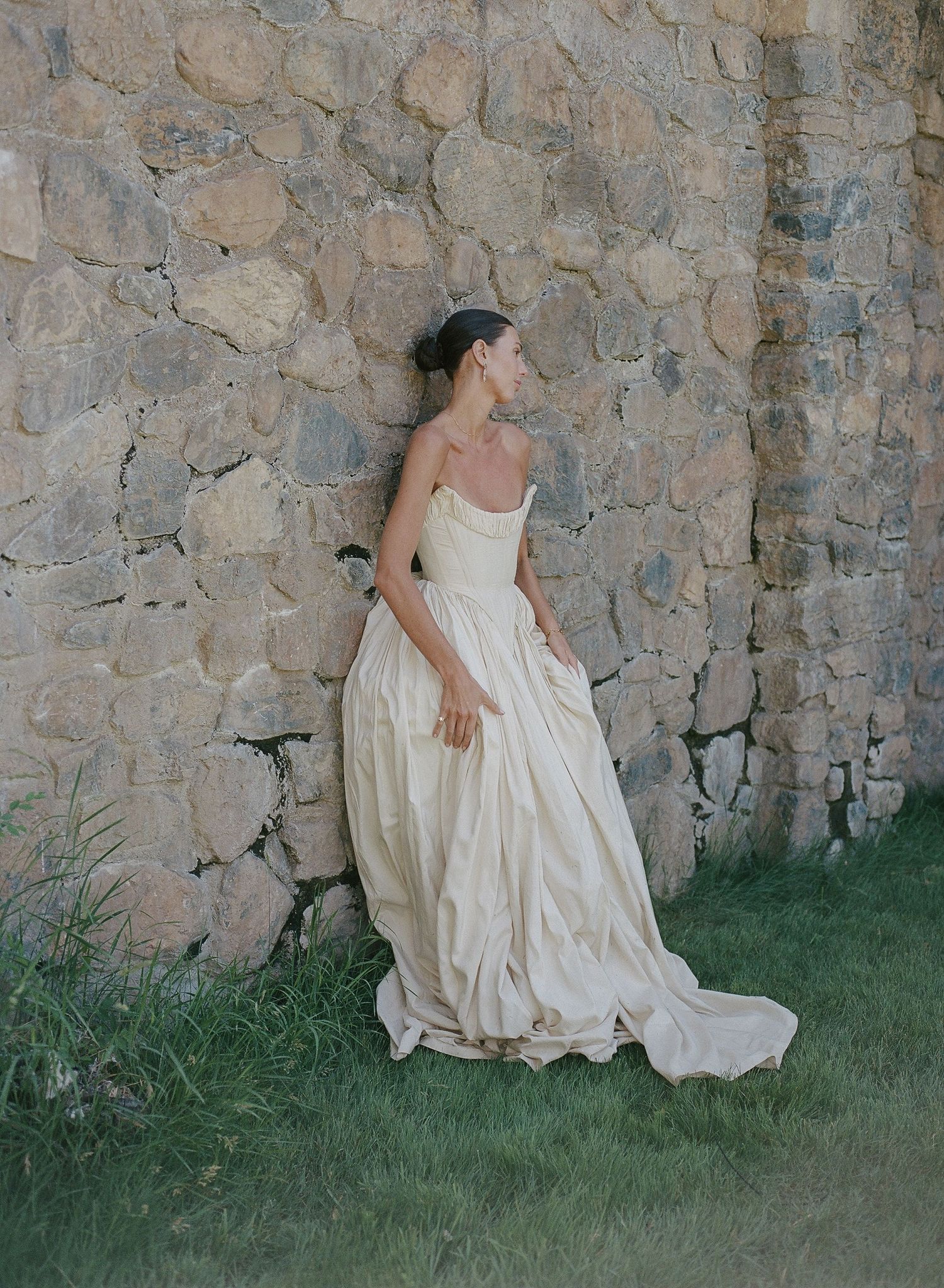 Bride in strapless ivory ballgown leans against rustic stone wall on green grass, gazing sideways in elegant outdoor setting.