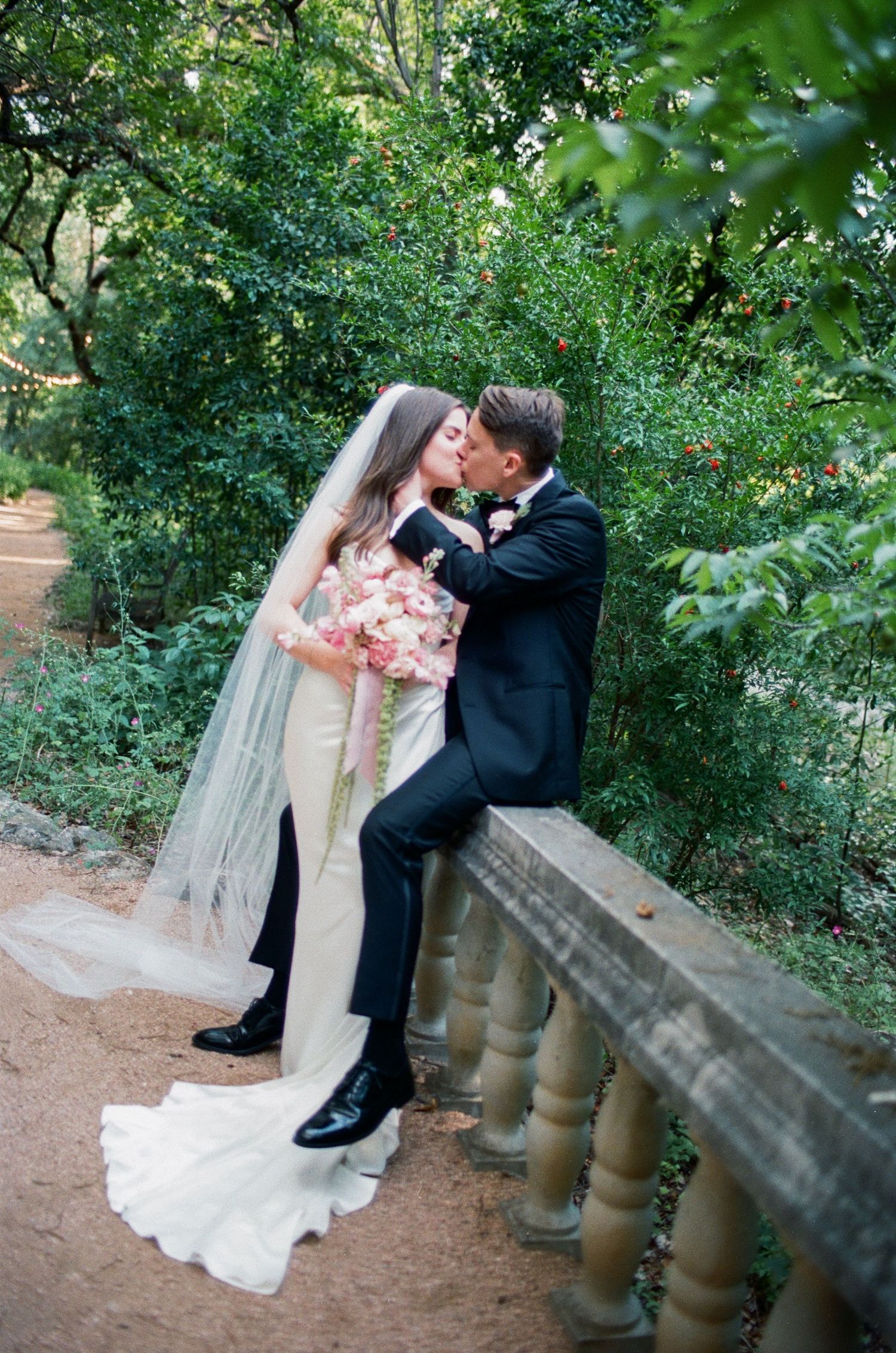 Romantic garden wedding portrait on stone railing surrounded by green foliage.