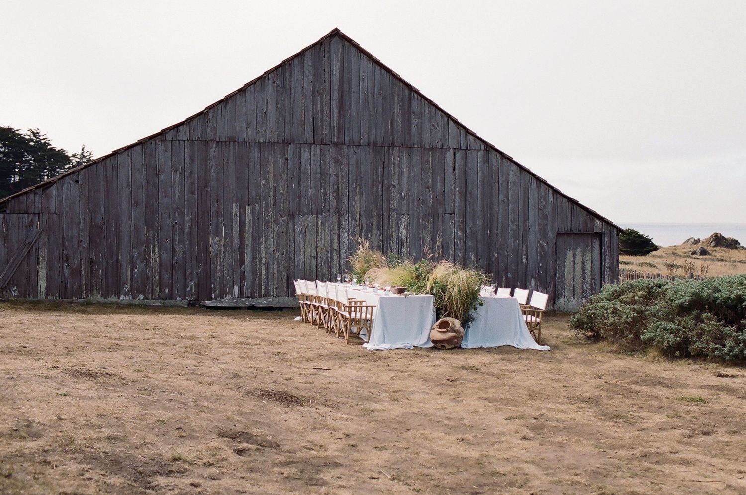 Rustic outdoor dining table set with white linens in front of a weathered gray barn on a dry field.