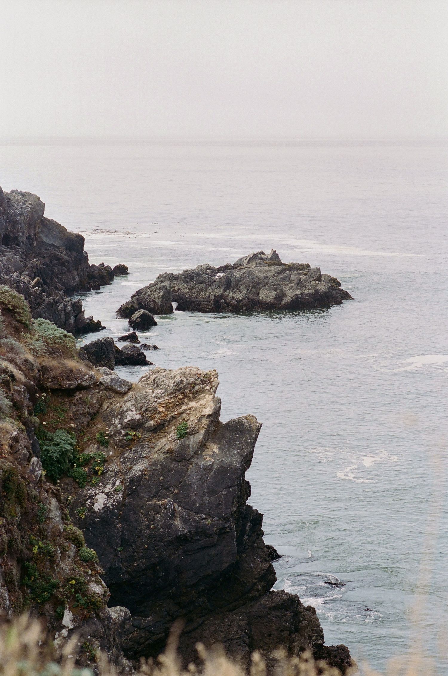 Rocky coastal cliffs overlooking calm ocean waters with misty horizon and small rock formations emerging from the sea.