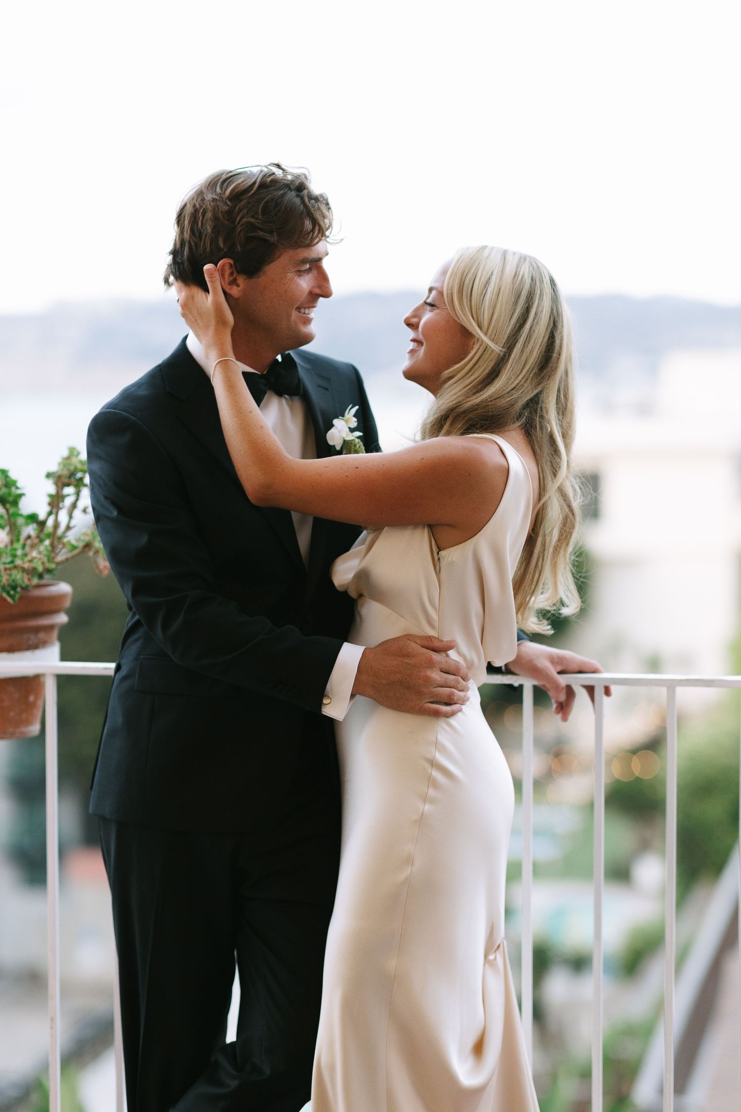 A romantic wedding couple embraces on a balcony, with the bride in a sleek white dress and groom in classic tuxedo.
