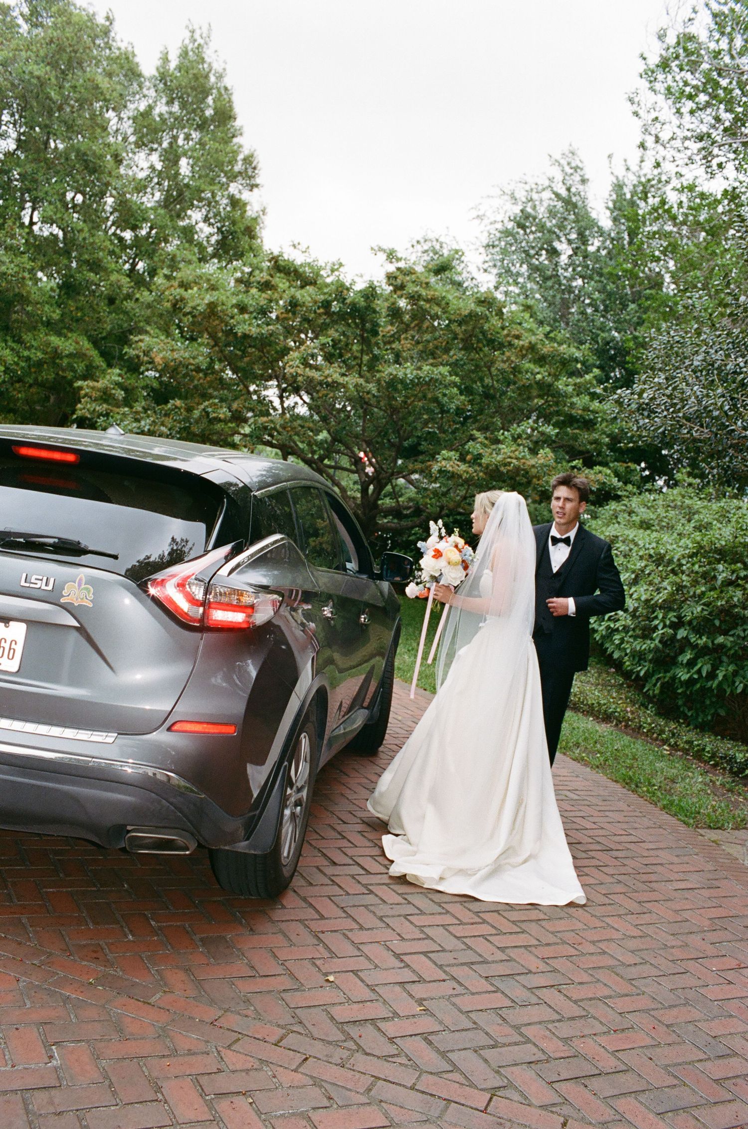 A bride and groom walk towards a gray car parked on brick pavement.