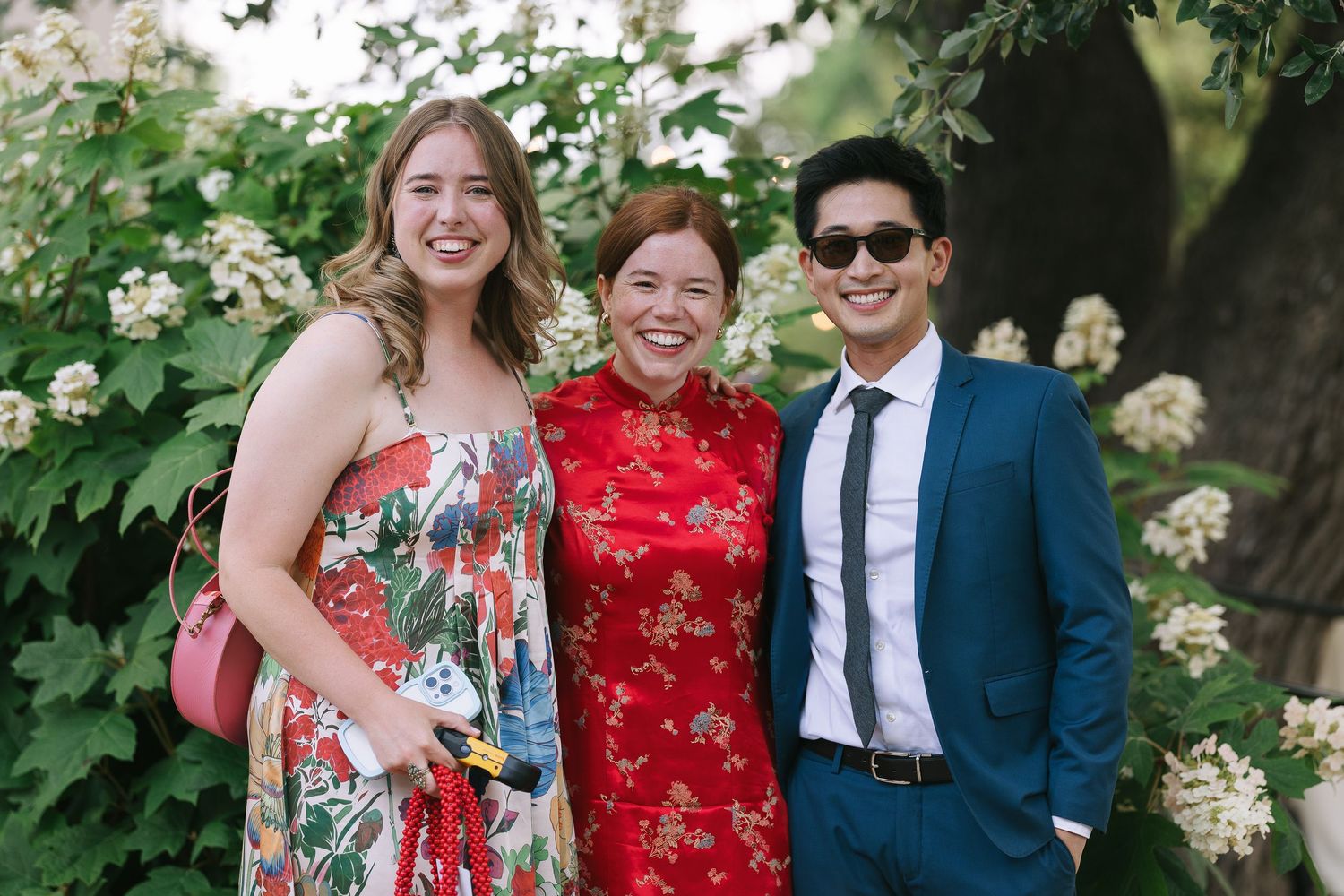 Wedding guests in floral dresses and formal attire gather outdoors near blooming white flowers.