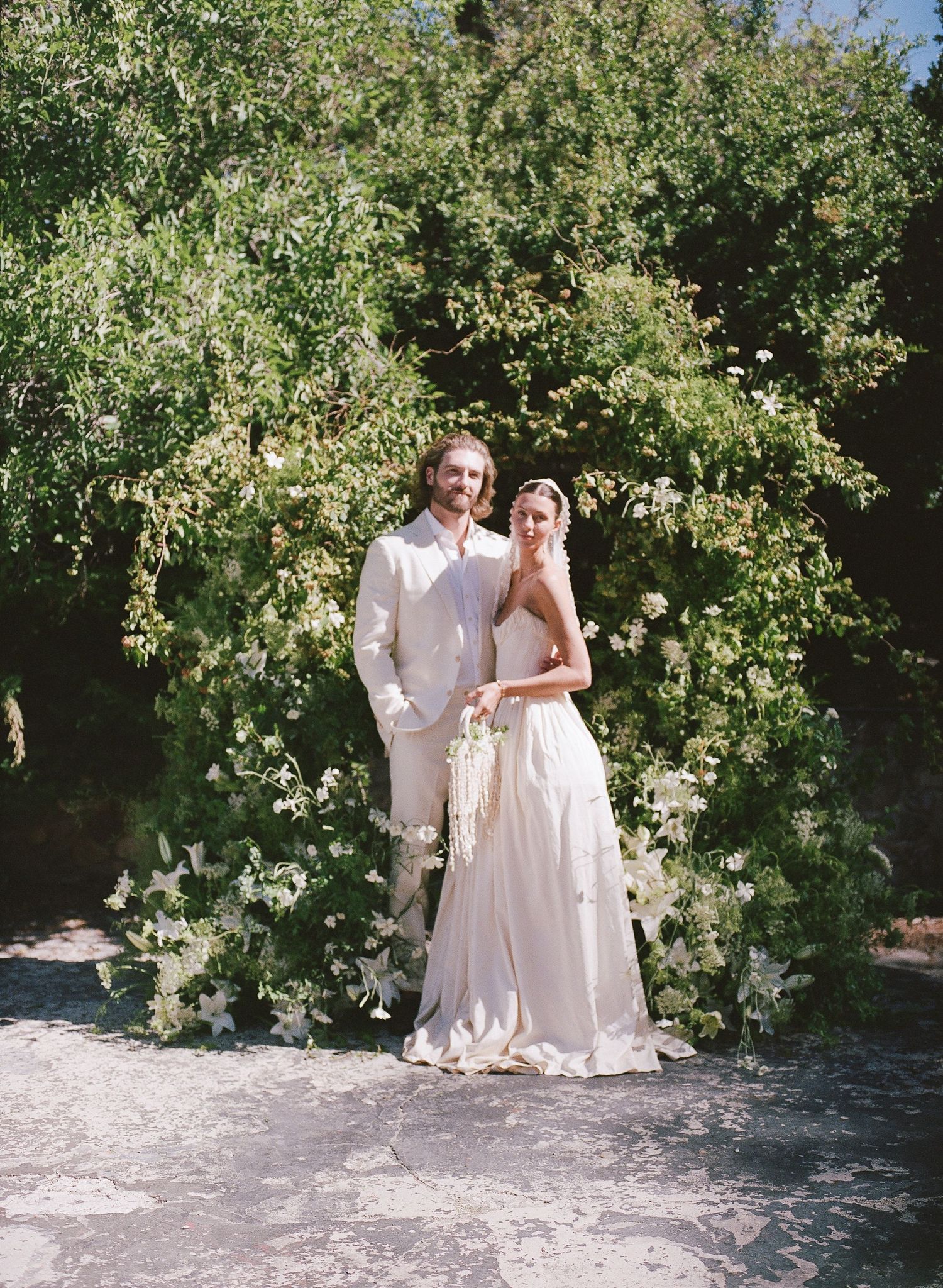 Bride and groom pose before lush green floral arch with white blooms at outdoor garden wedding ceremony.