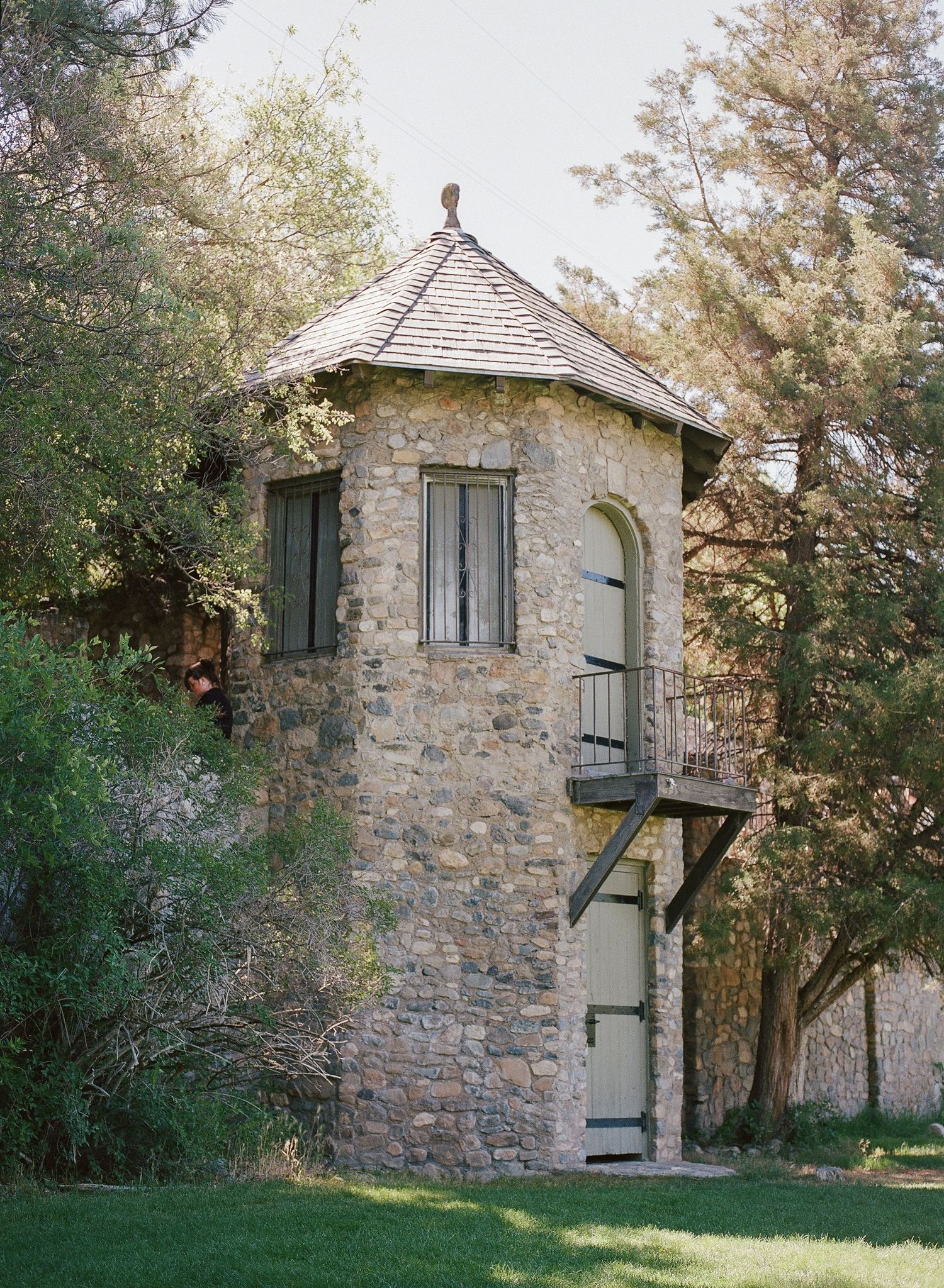 Rustic stone tower with conical roof, arched window, and spiral staircase surrounded by lush green trees.