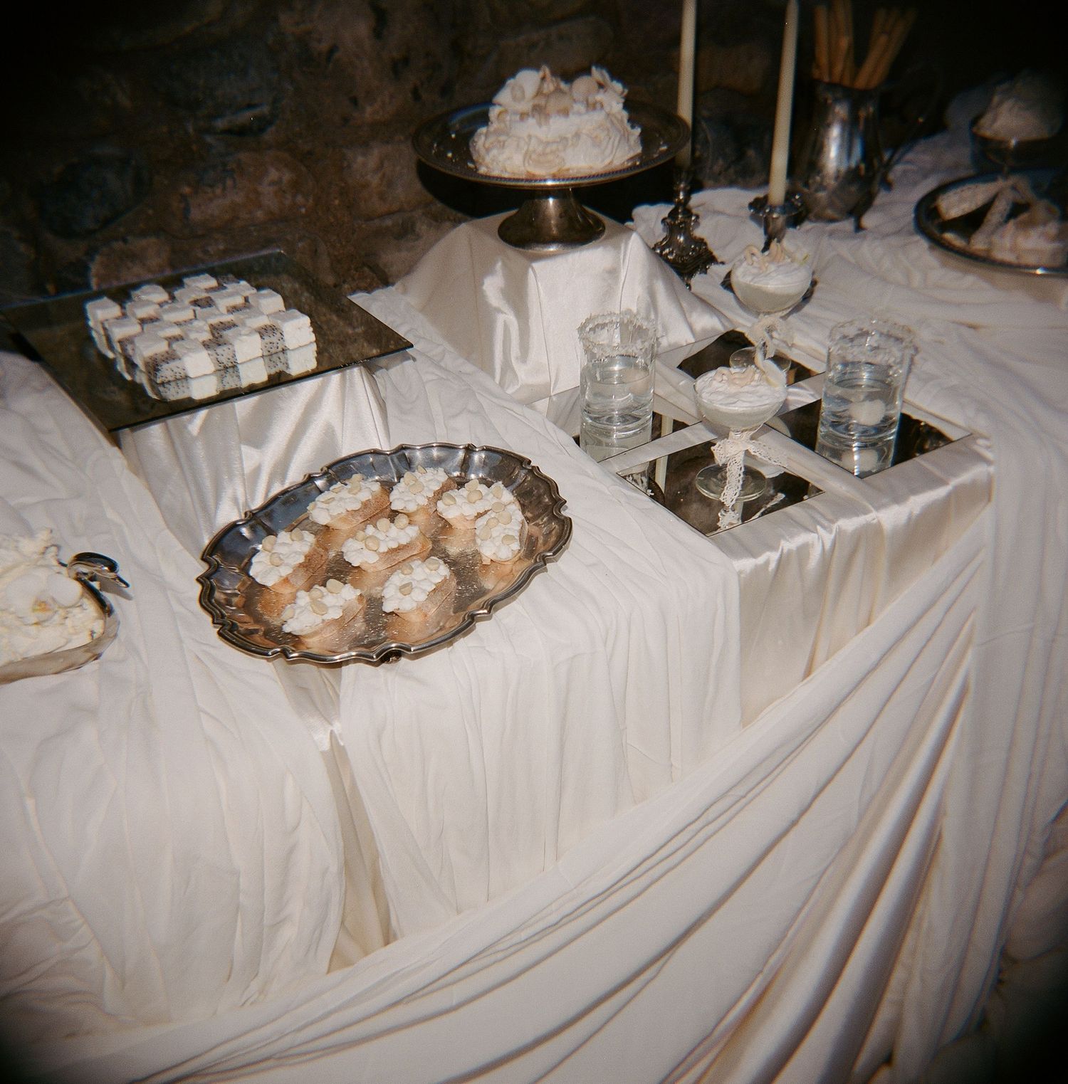 Elegant white draped dessert table with silver platters of treats, candles, and glassware at a formal event.