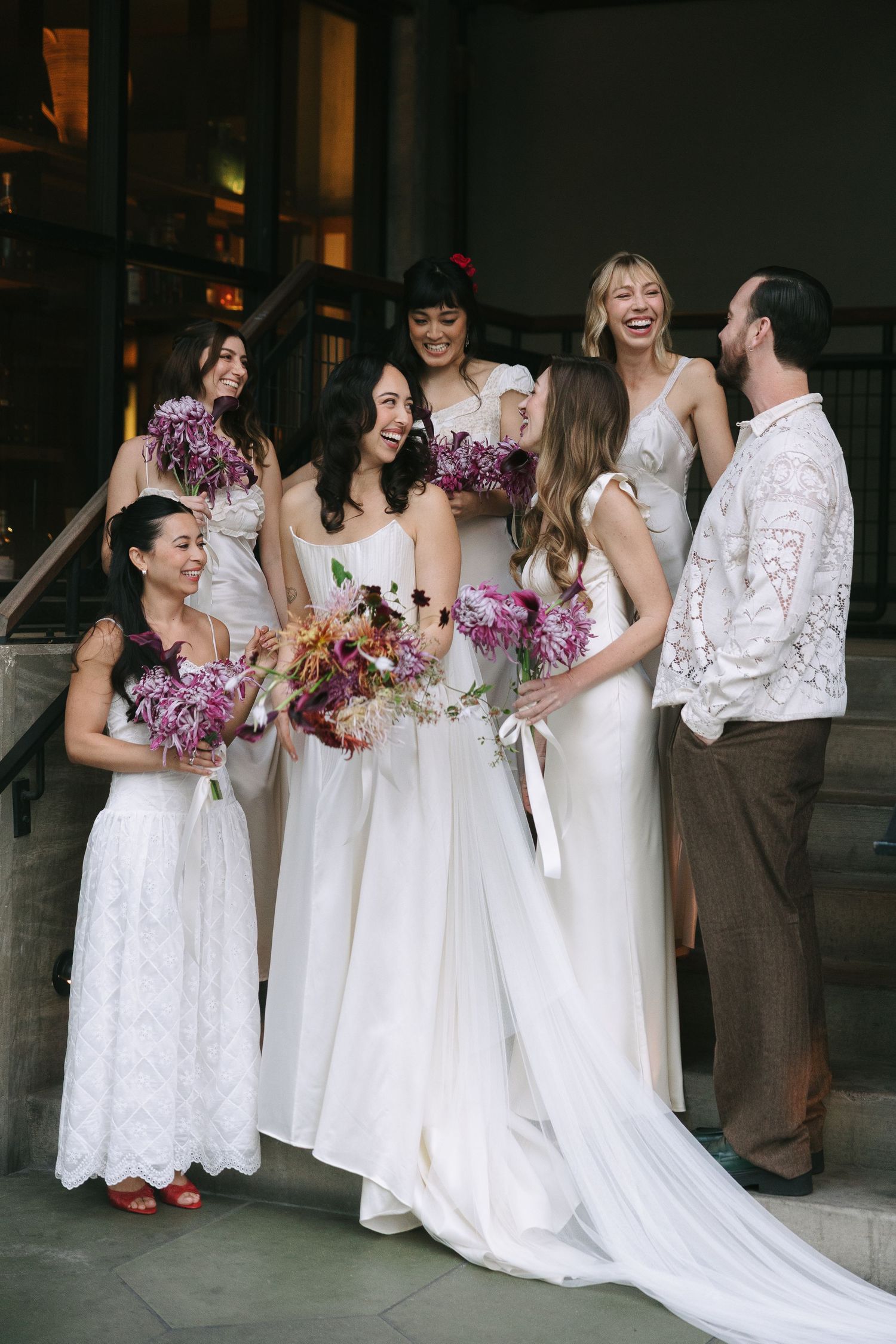 Group of bridesmaids in white lace dresses laugh together while holding purple floral arrangements.