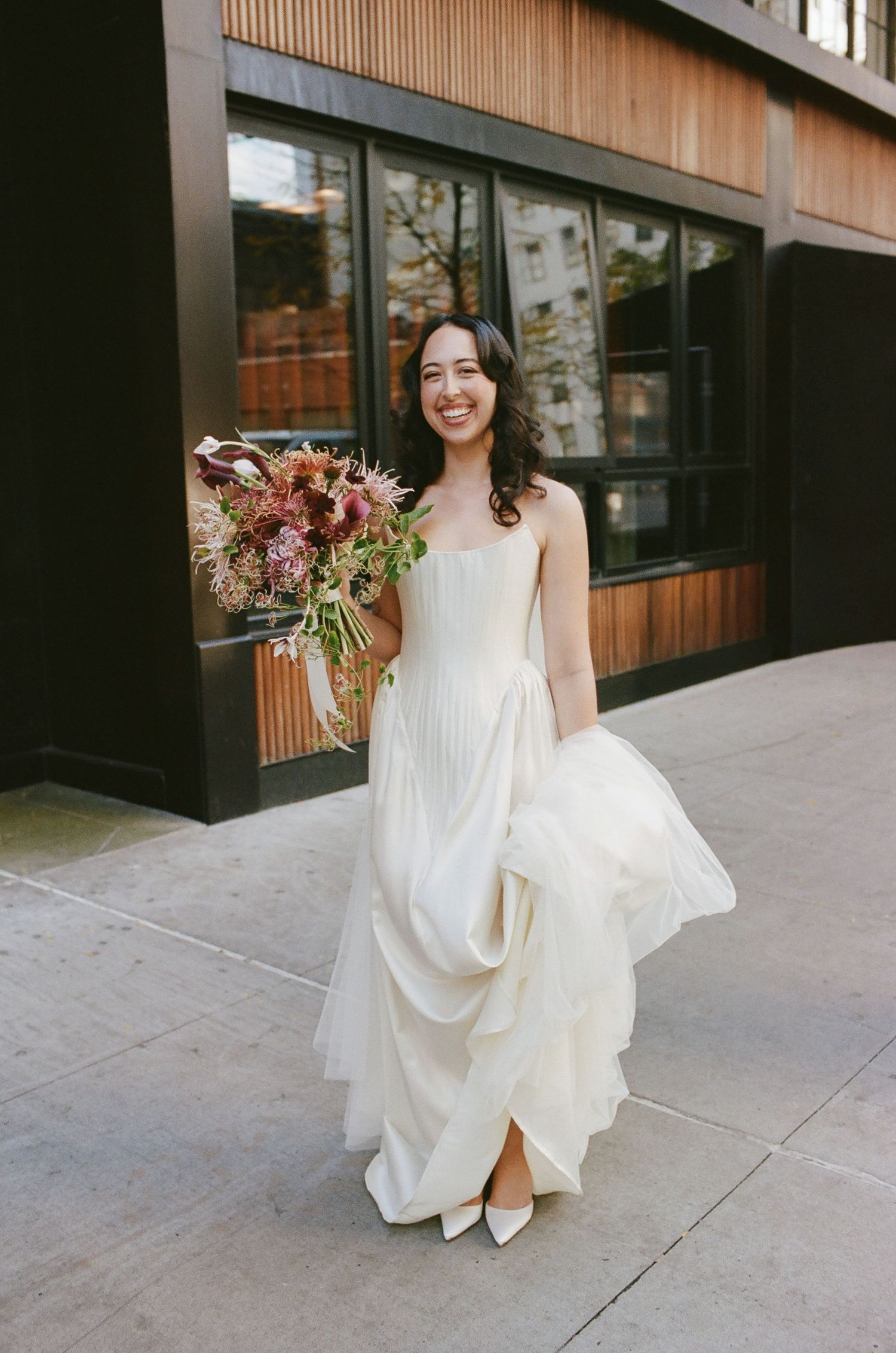 A bride in a flowing white dress holds a vibrant autumn bouquet while posing on a city sidewalk.