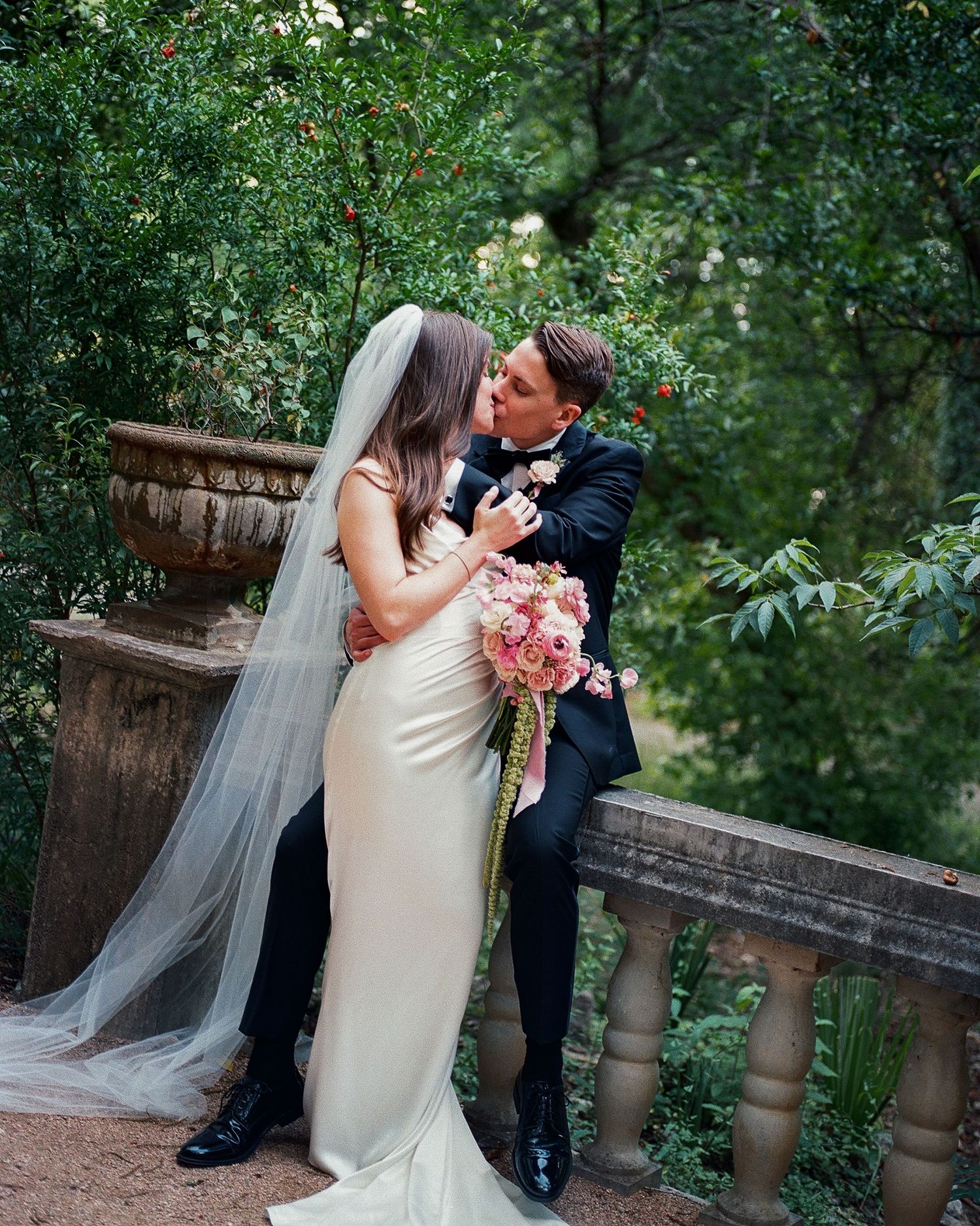 Wedding couple embracing in an elegant garden setting with stone architecture.