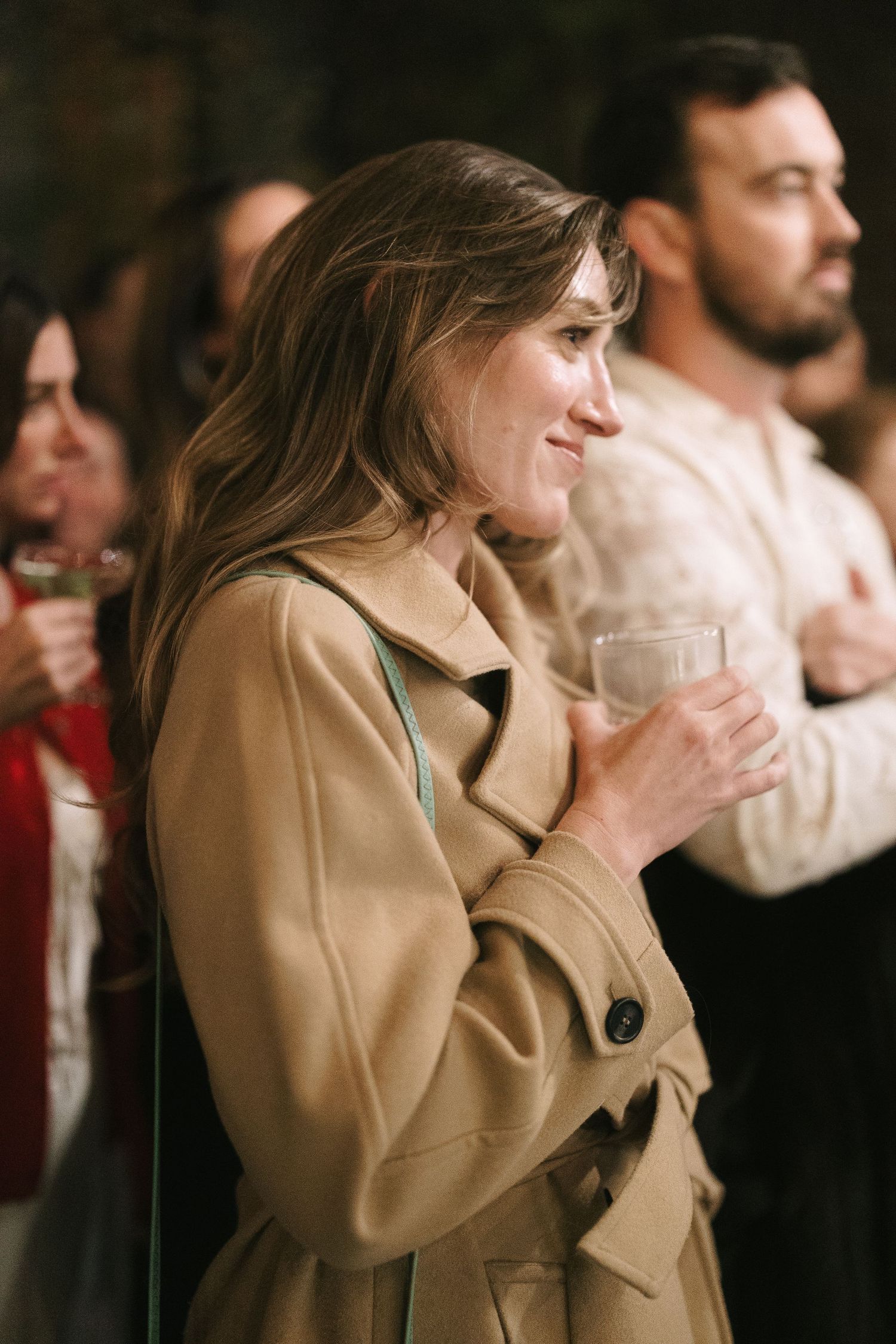 Guest in tan trench coat smiling while holding wine glass at evening event.
