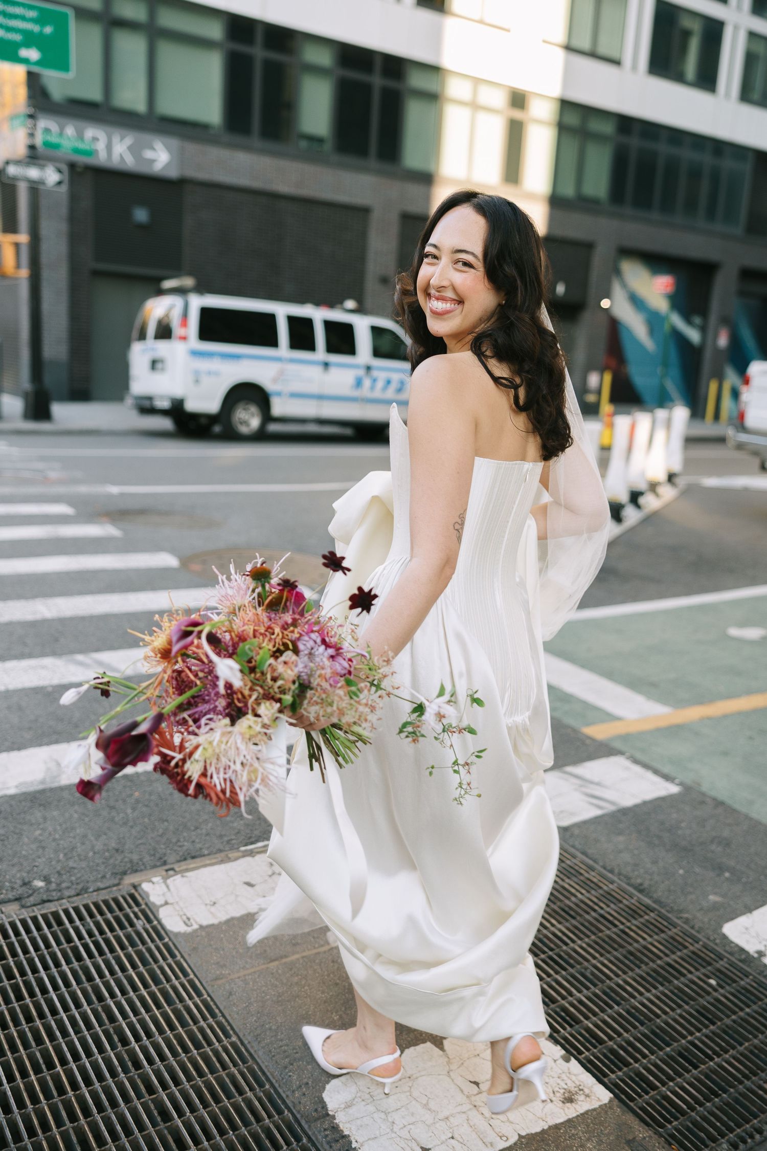 A bride twirls playfully on a city sidewalk grate while holding her floral bouquet.