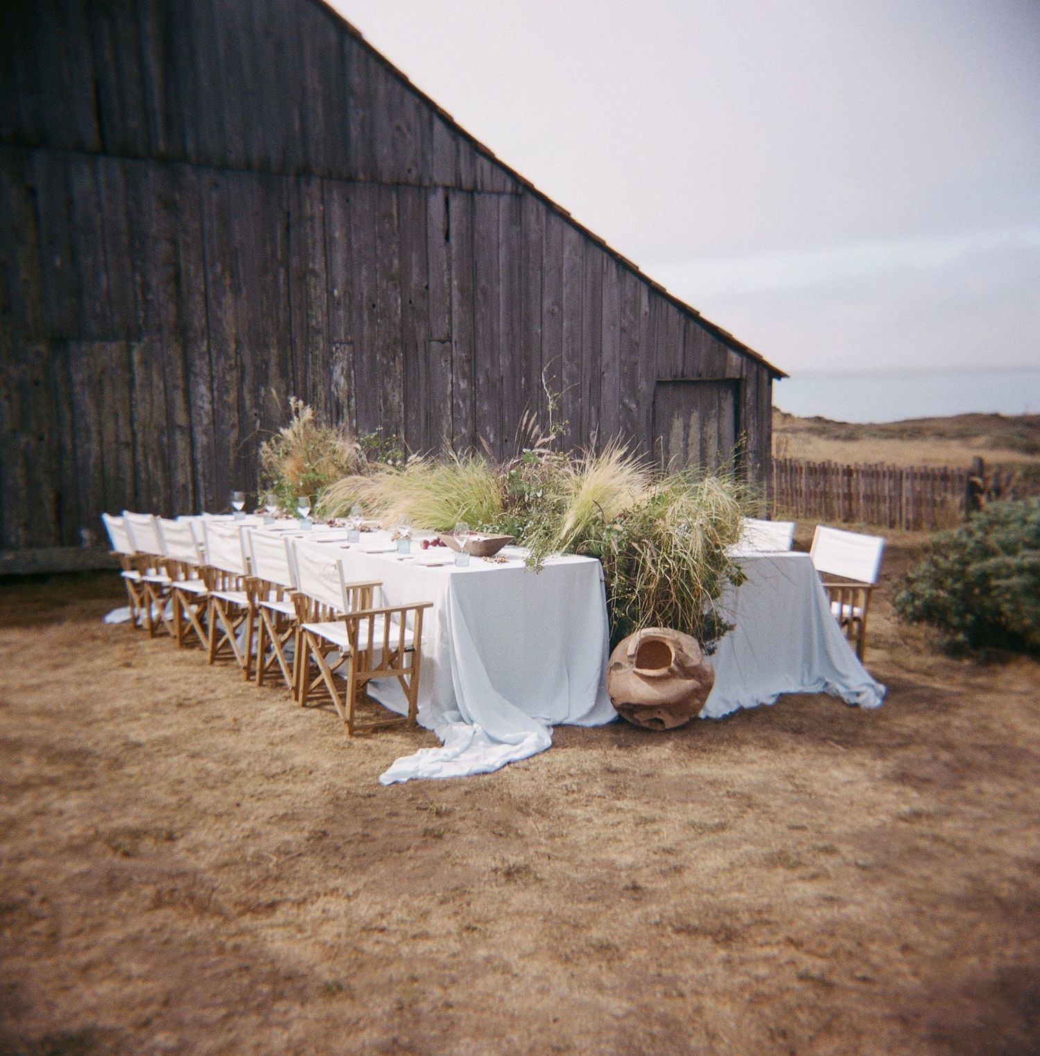 Rustic outdoor wedding reception setup with wooden chairs and white linens beside a weathered barn.